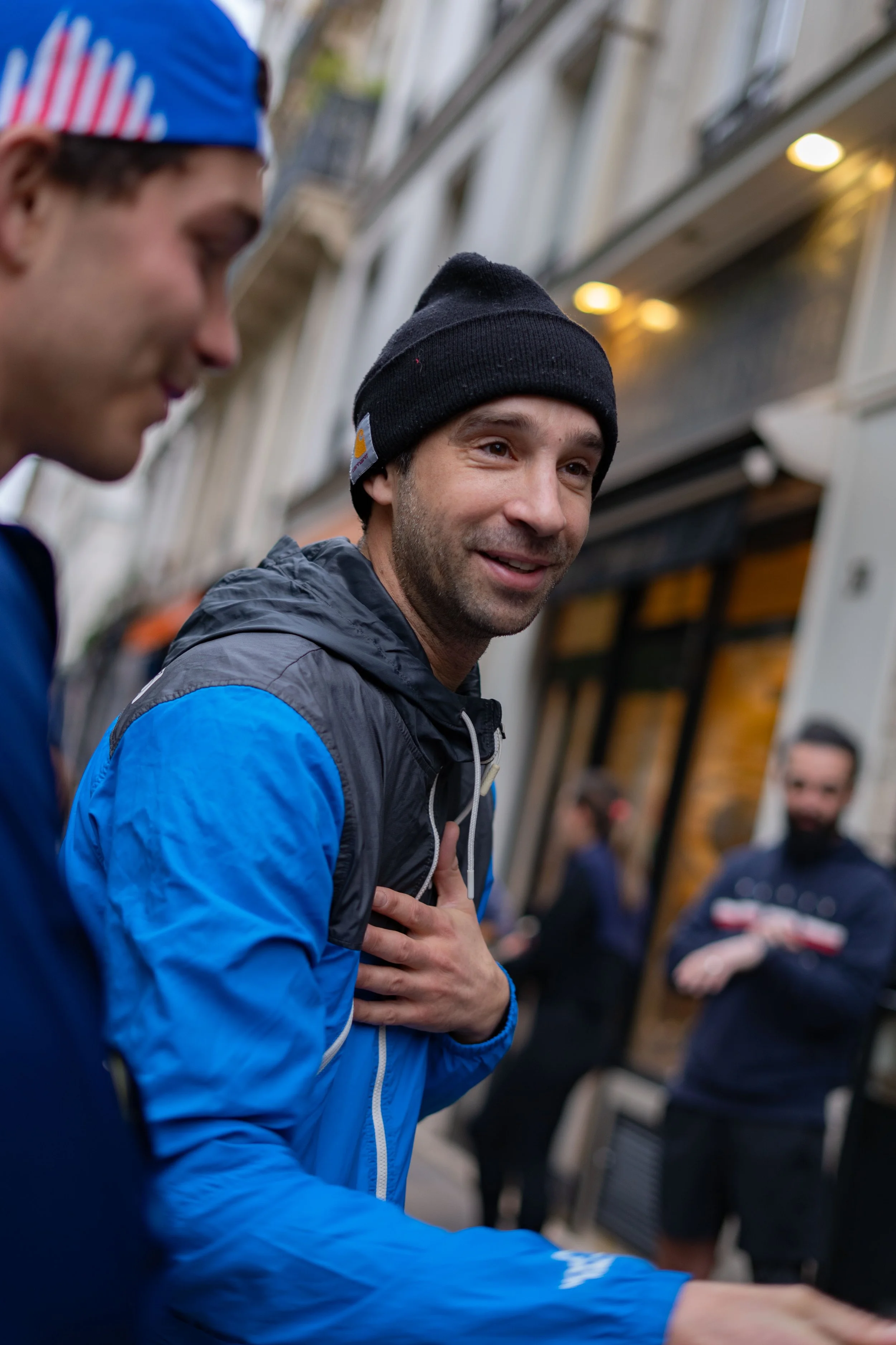 Un groupe de personnes, principalement des hommes, discutent dans une rue urbaine. L'homme au centre porte un bonnet noir et une veste bleue, sourit, et semble impliqué dans une conversation amicale.