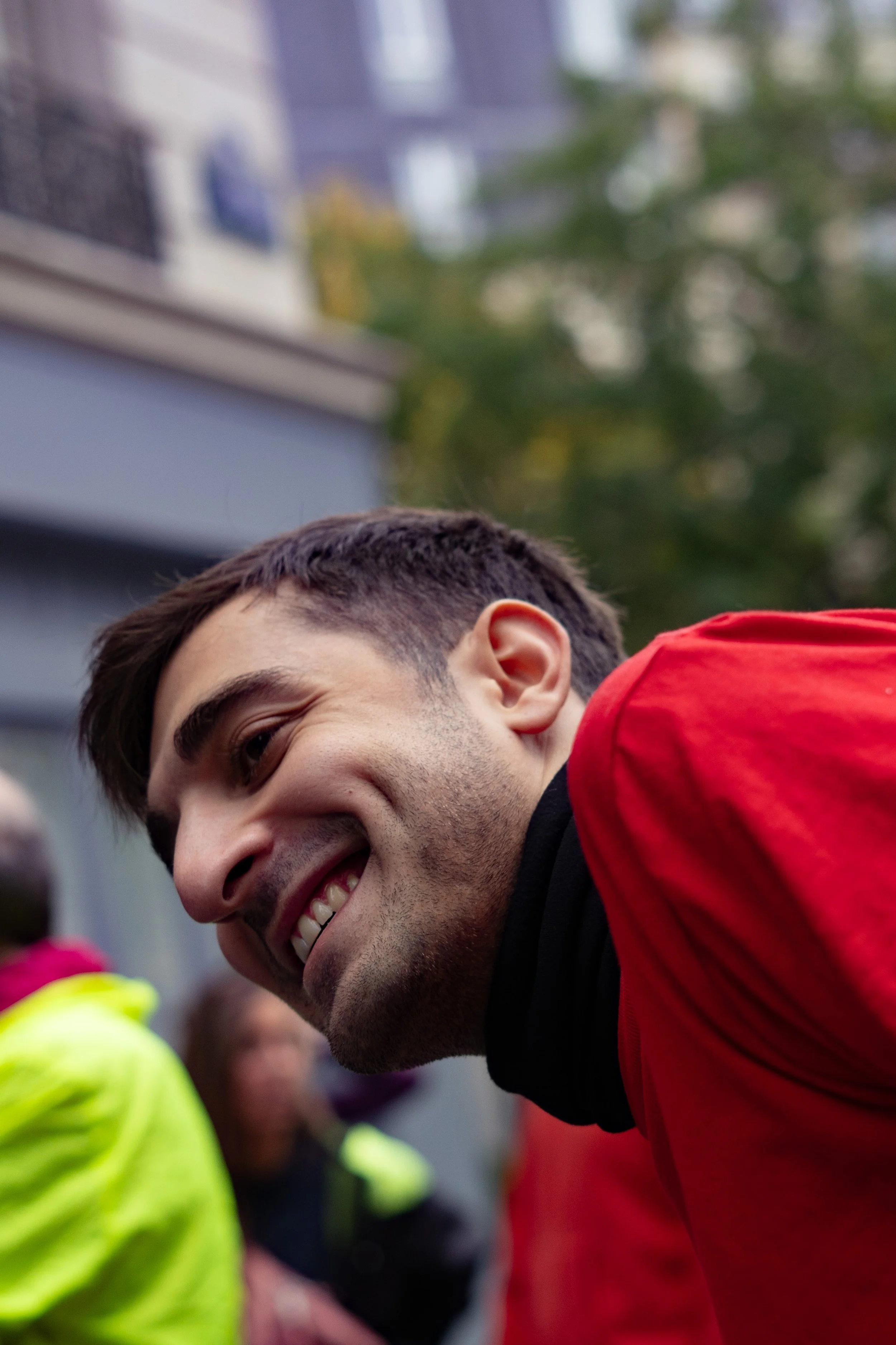 Jeune homme souriant, porteur d'une veste rouge, en interaction avec des personnes en tenues de police ou de sécurité, lors d'un événement public, en extérieur avec des bâtiments en arrière-plan.
