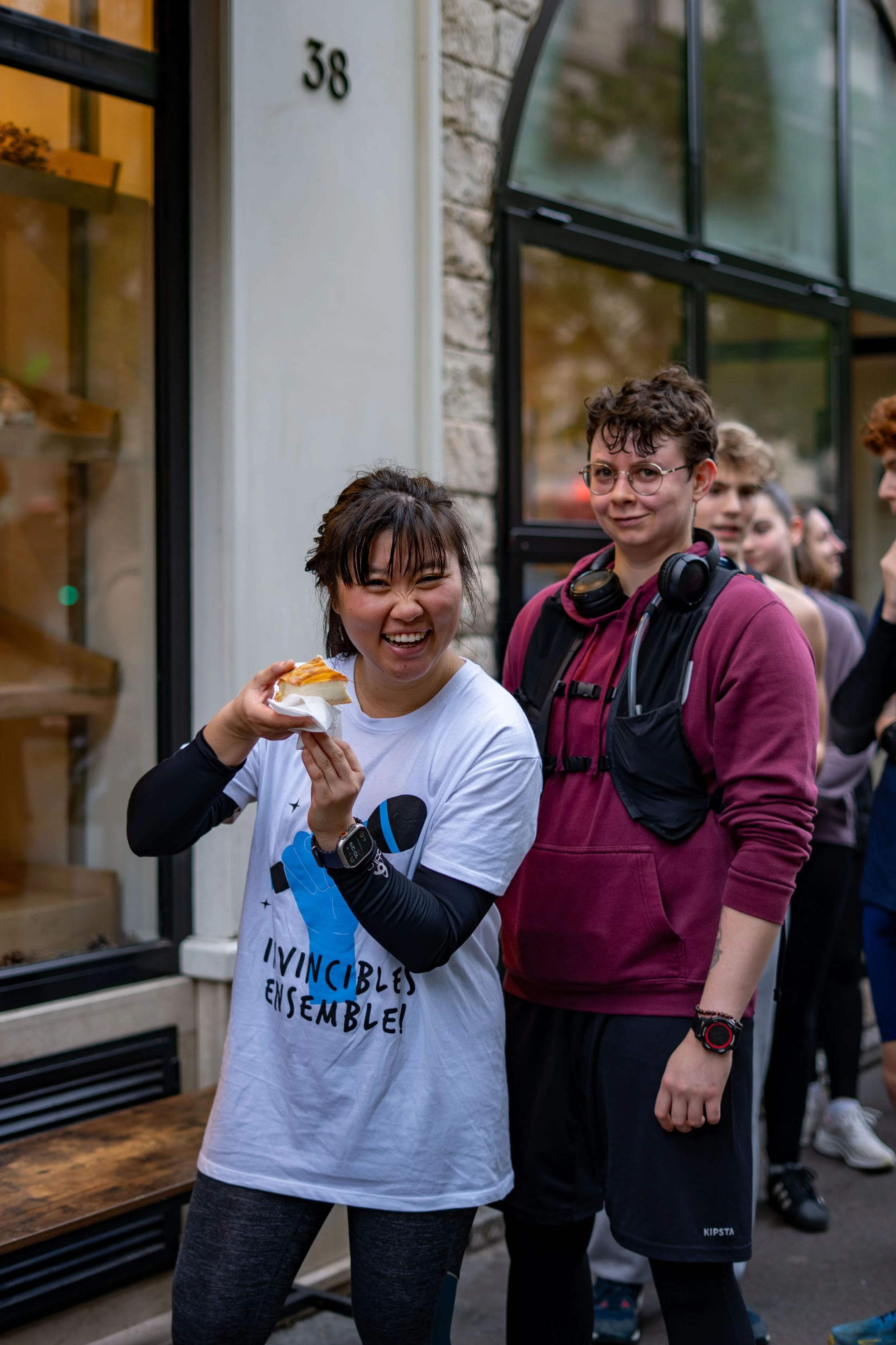 Groupe de jeunes souriants dehors, une fille tient une part de tarte, en arrière-plan un bâtiment avec un numéro 38.