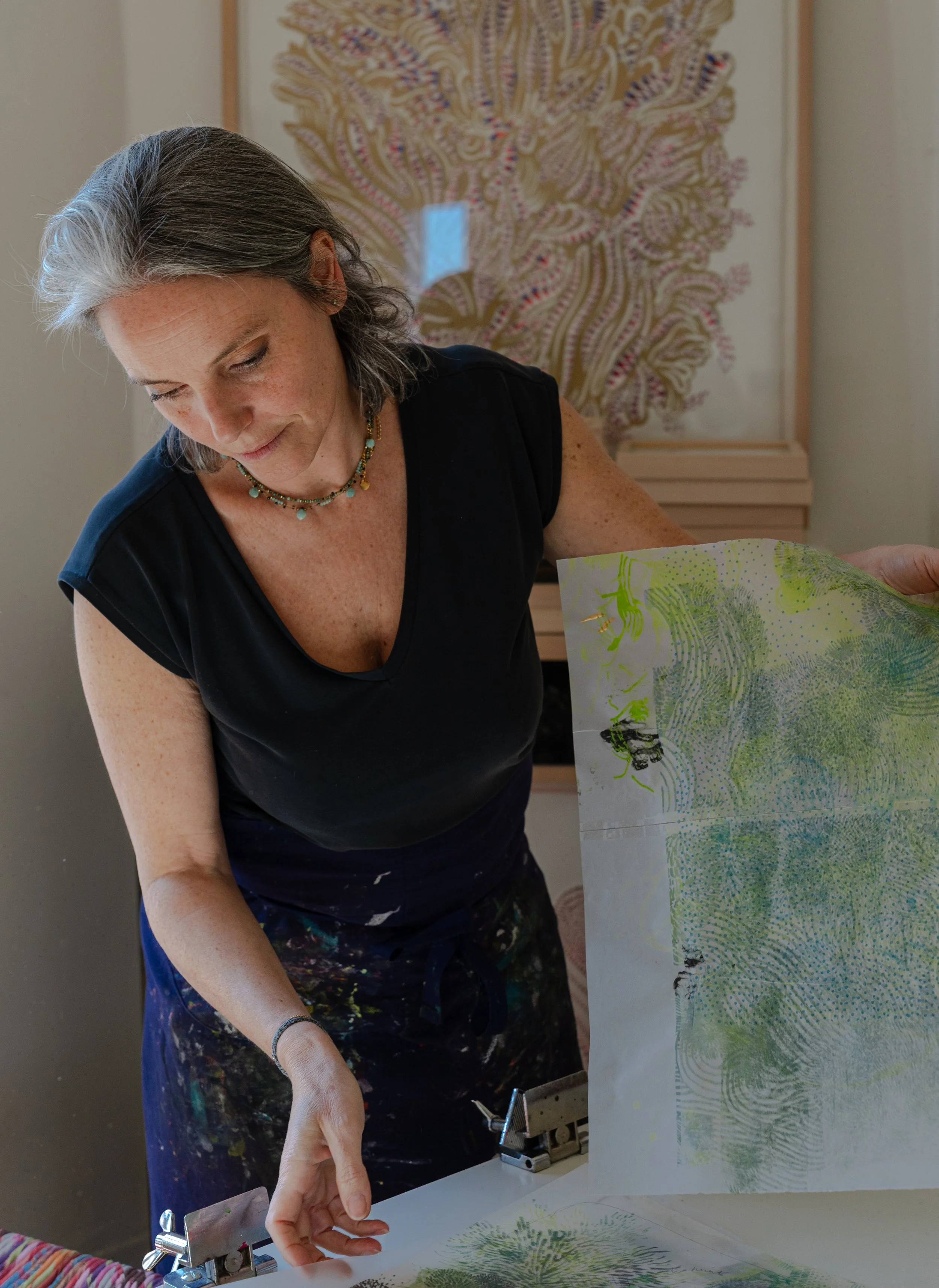 Une femme âgée avec des cheveux gris, portant un collier coloré, qui travaille sur une œuvre d'art en utilisant une presse à imprimer, avec une grande feuille de papier imprimée de motifs d'arbres et de paysages colorés.