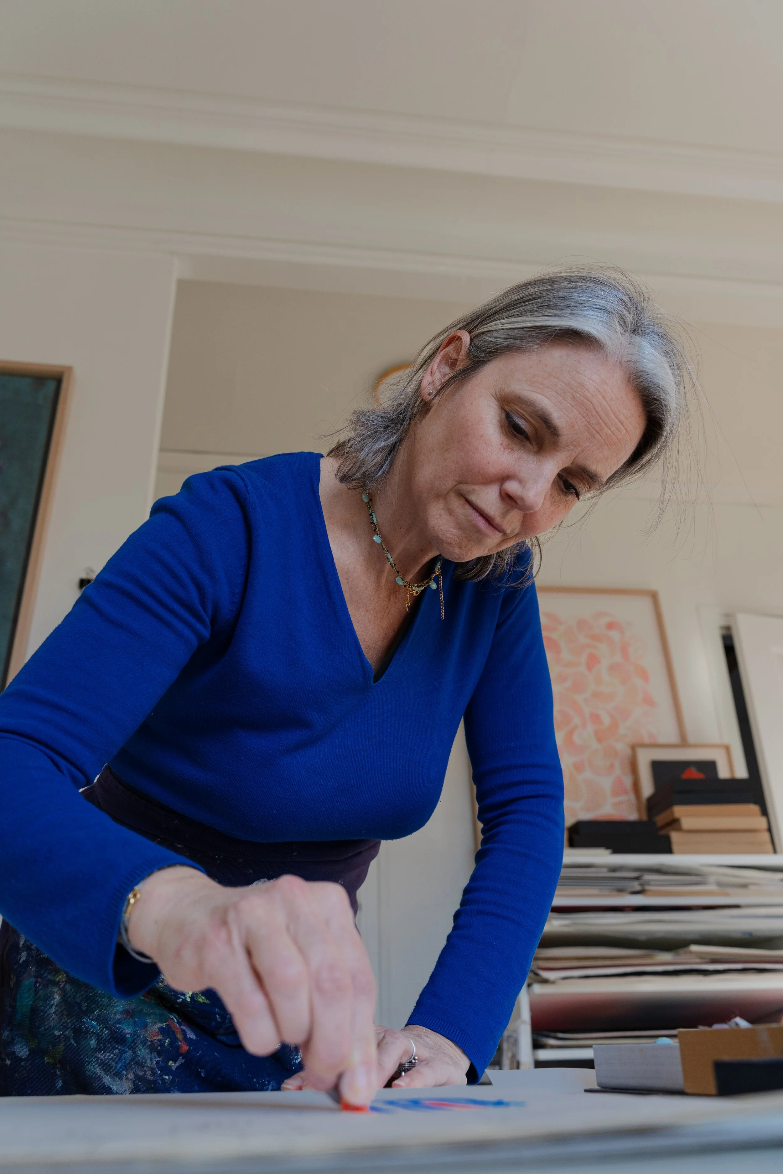 Une femme avec des cheveux gris porteur d'une chemise bleue travaille sur un projet avec des papiers et des crayons dans un espace intérieur.