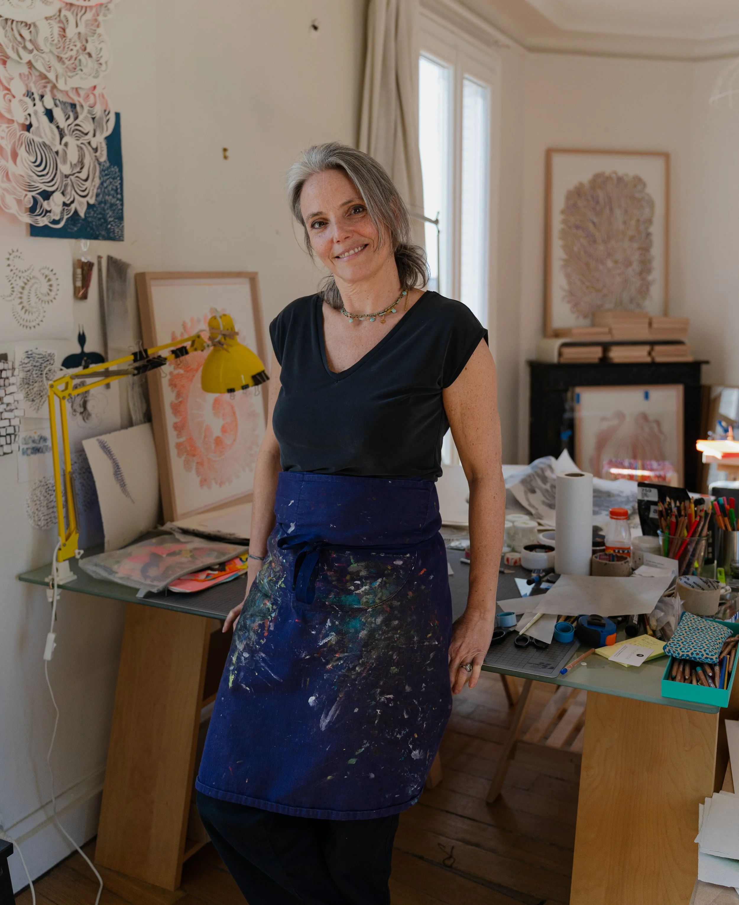 Une femme souriante dans un atelier d'art, portant un tablier de peinture avec des taches de peinture, entourée d'œuvres d'art et de fournitures artistiques.