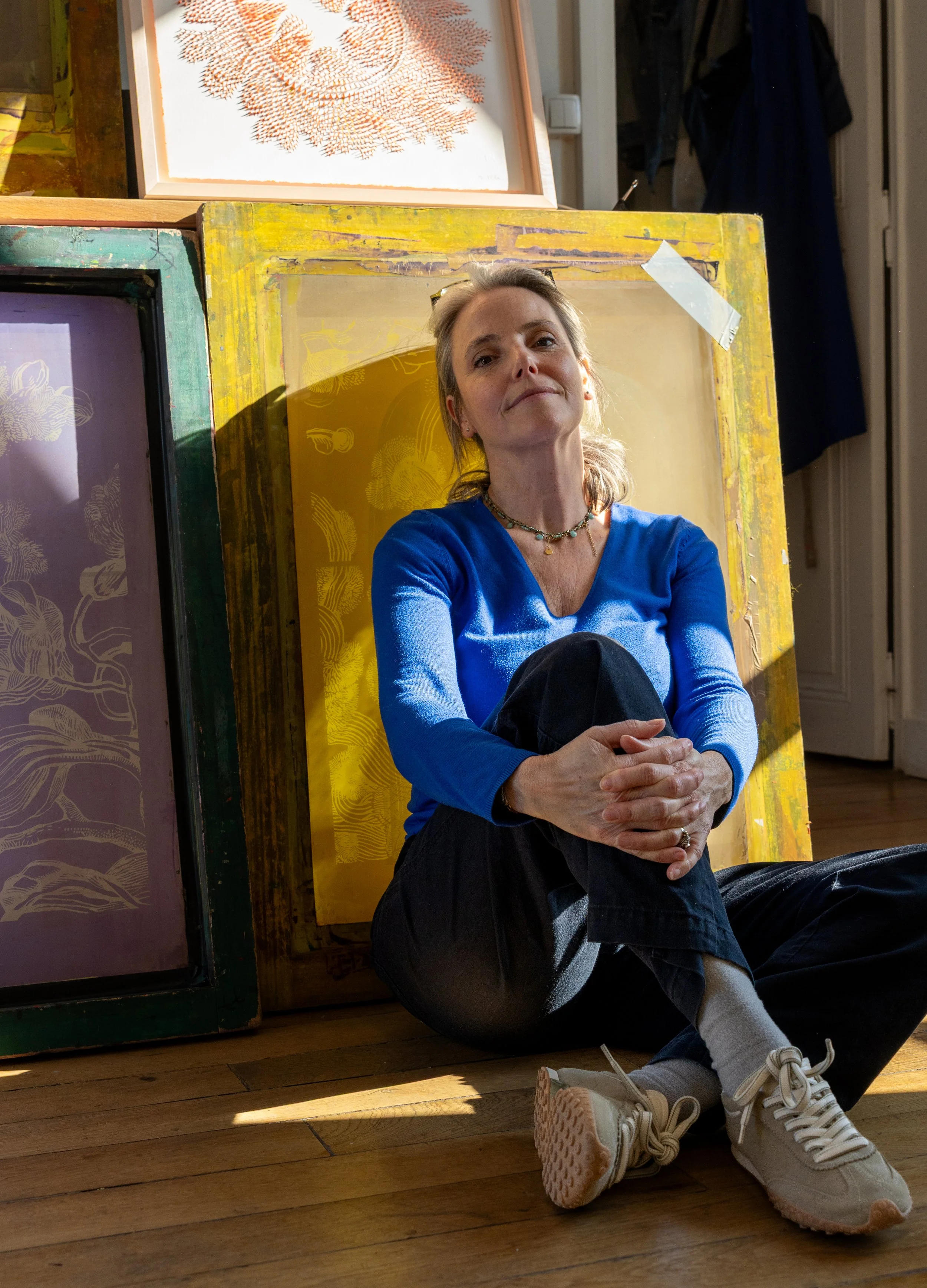 Une femme assise par terre devant des meubles colorés, portant un pull bleu, des pantalons noirs, des chaussettes blanches et des baskets beige, avec des œuvres d'art en arrière-plan.