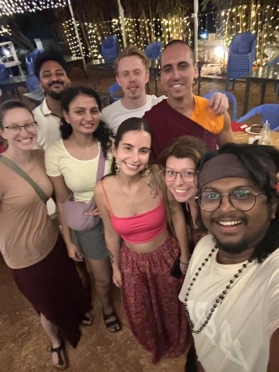 Group of people smiling for a selfie at a nighttime outdoor gathering with string lights and blue chairs in the background.