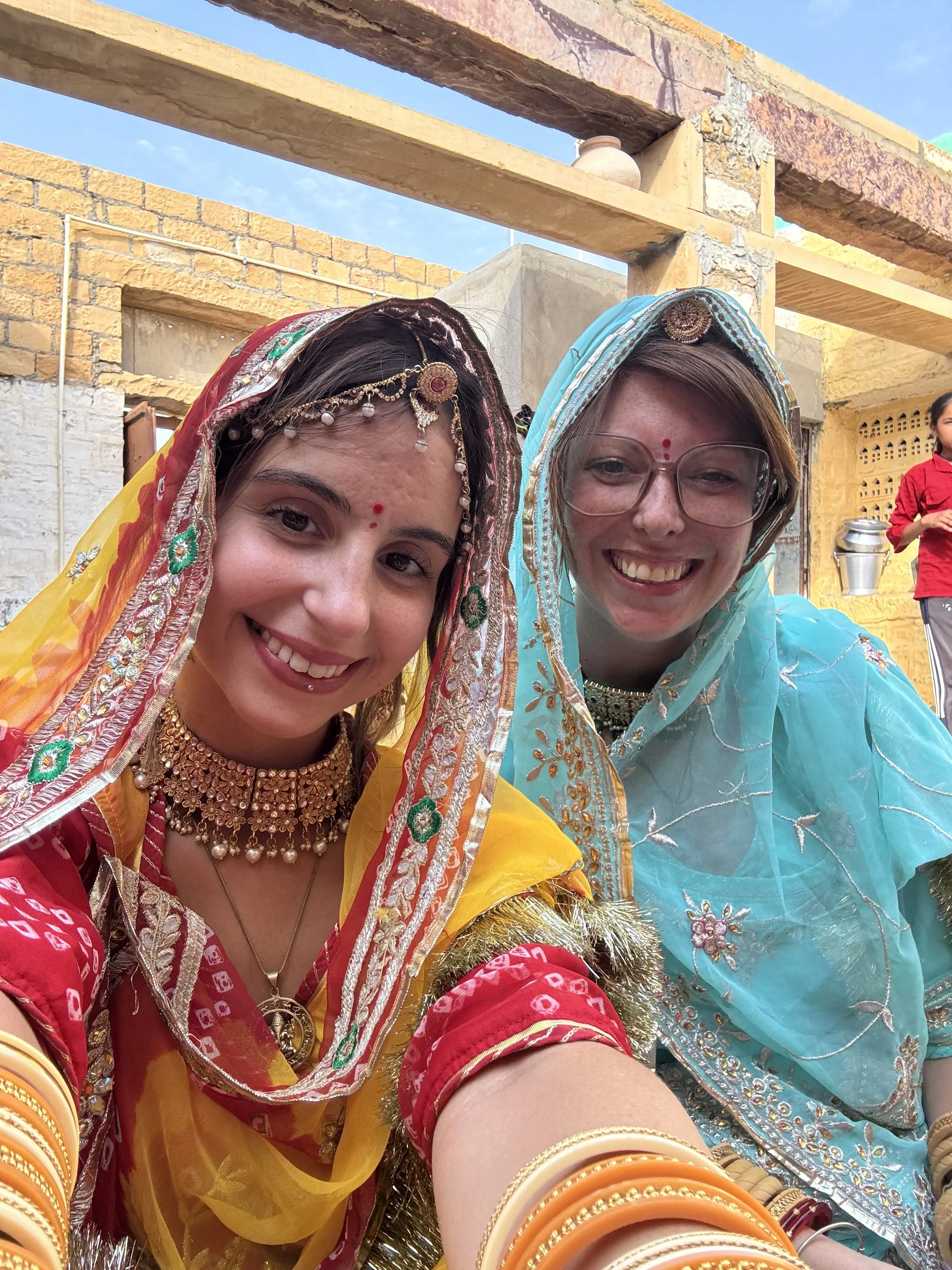 Two women dressed in traditional Indian attire, smiling for a selfie at an outdoor event with brick walls and a blue sky in the background.
