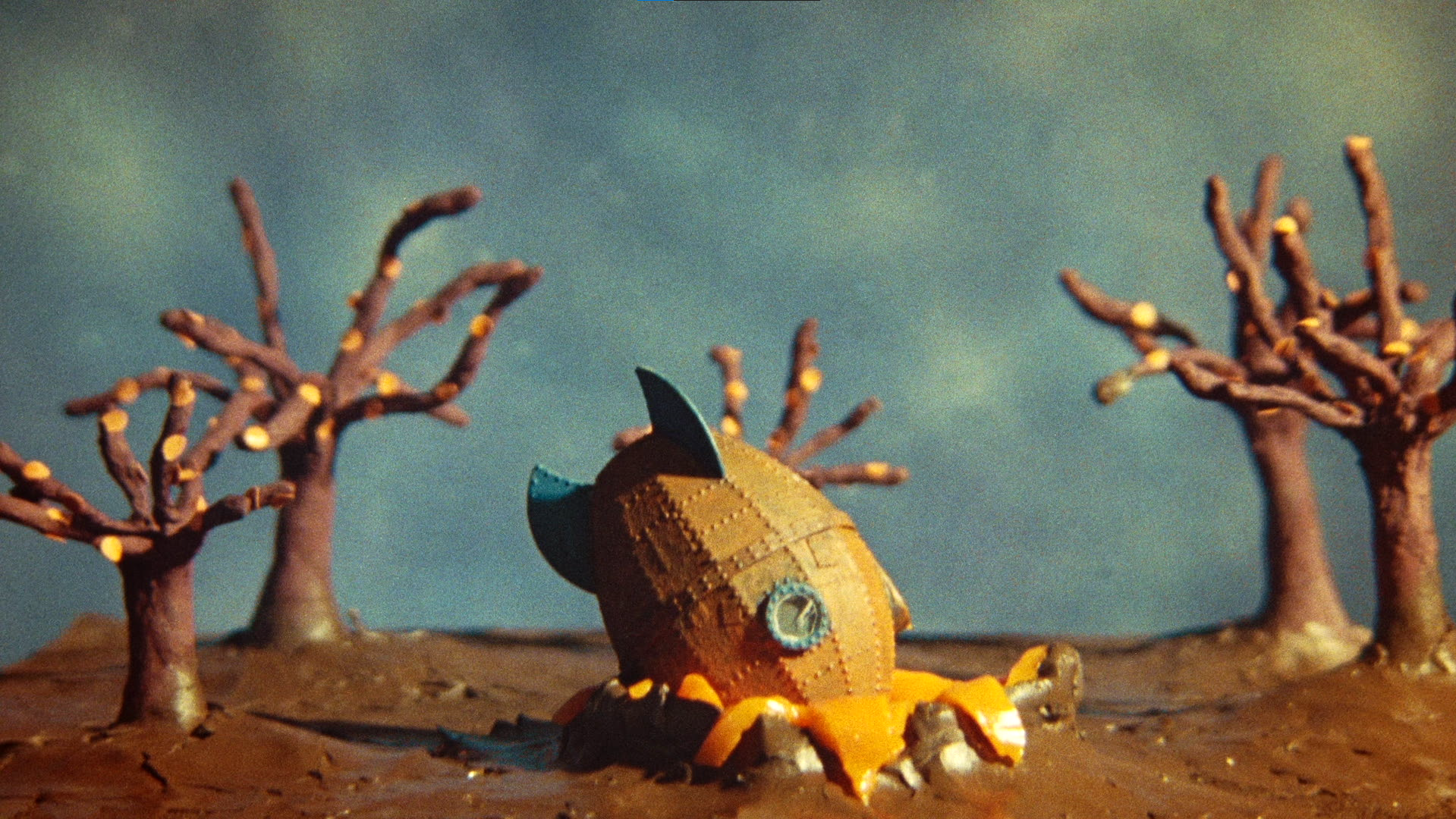 A clay or papier-mâché model of a rocket ship sits on the ground among small trees with painted lights, under a cloudy sky.
