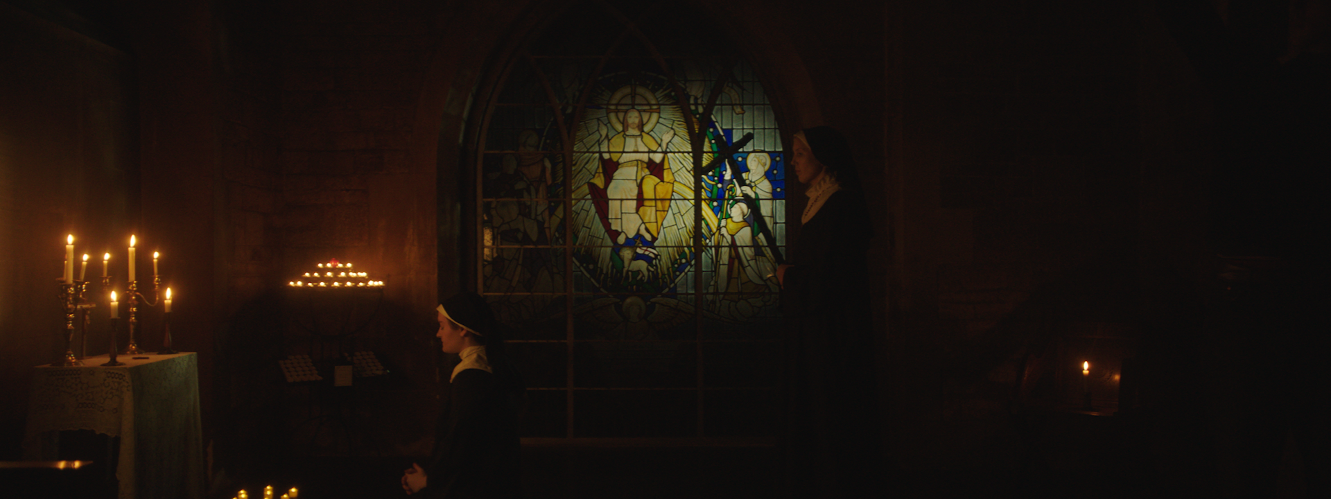 A dimly lit church interior with a stained glass window depicting Jesus and angels. Two nuns, one adult and one child, are praying near a lit candle stand.