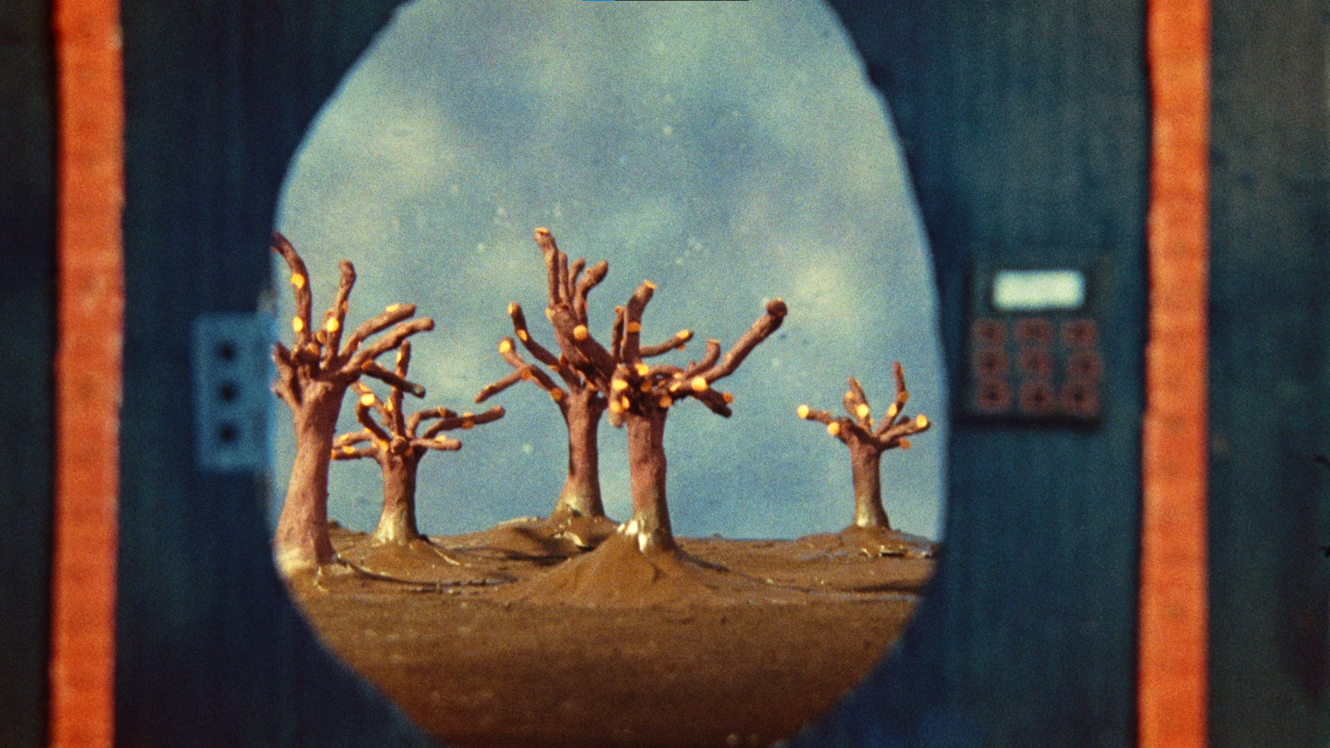 A miniature landscape model seen through the round opening of a small structure. It features small trees with bare branches, dry soil, and a cloudy sky background.