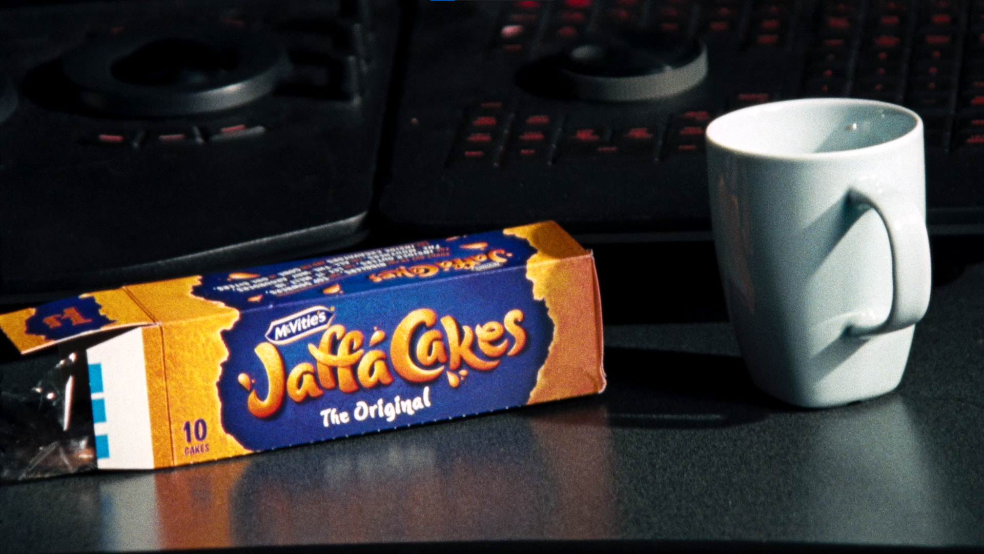 A box of McVitie's Jaffa Cakes and a white mug on a dark desk, with a computer keyboard and speaker in the background.