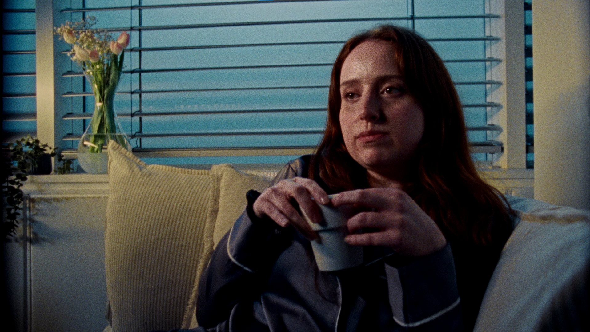 A woman with red hair sitting on a couch, holding a cup, in a room with blue window shutters and a vase of flowers on the window sill.