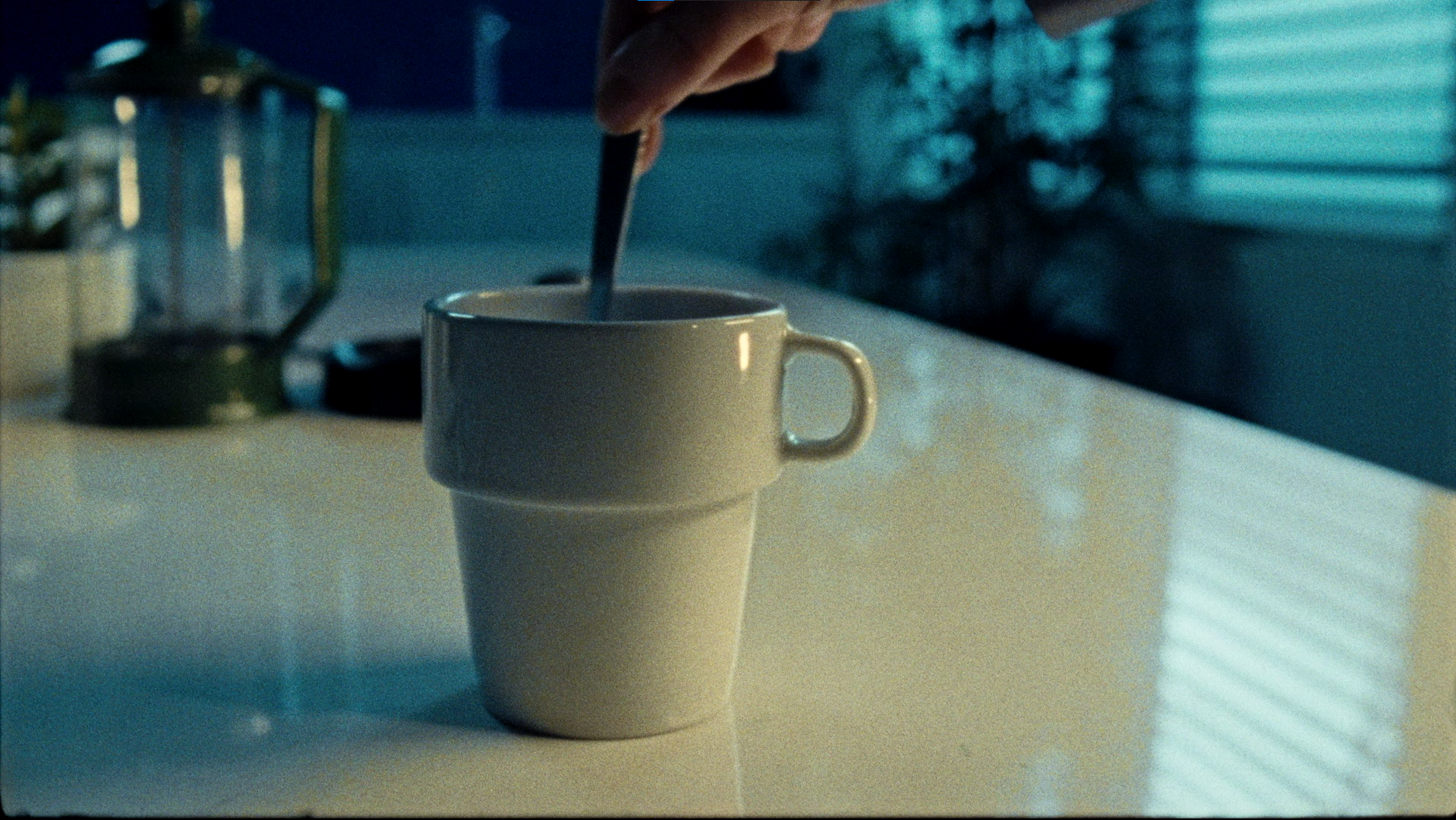 A white ceramic mug on a table with a person stirring a beverage inside, background with an out-of-focus lantern and window blinds.