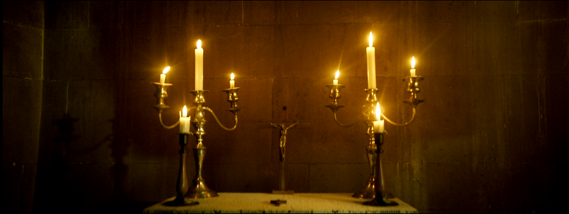 A religious altar with a cross and seven lit white candles in brass candle holders, set against a plain dark wall.