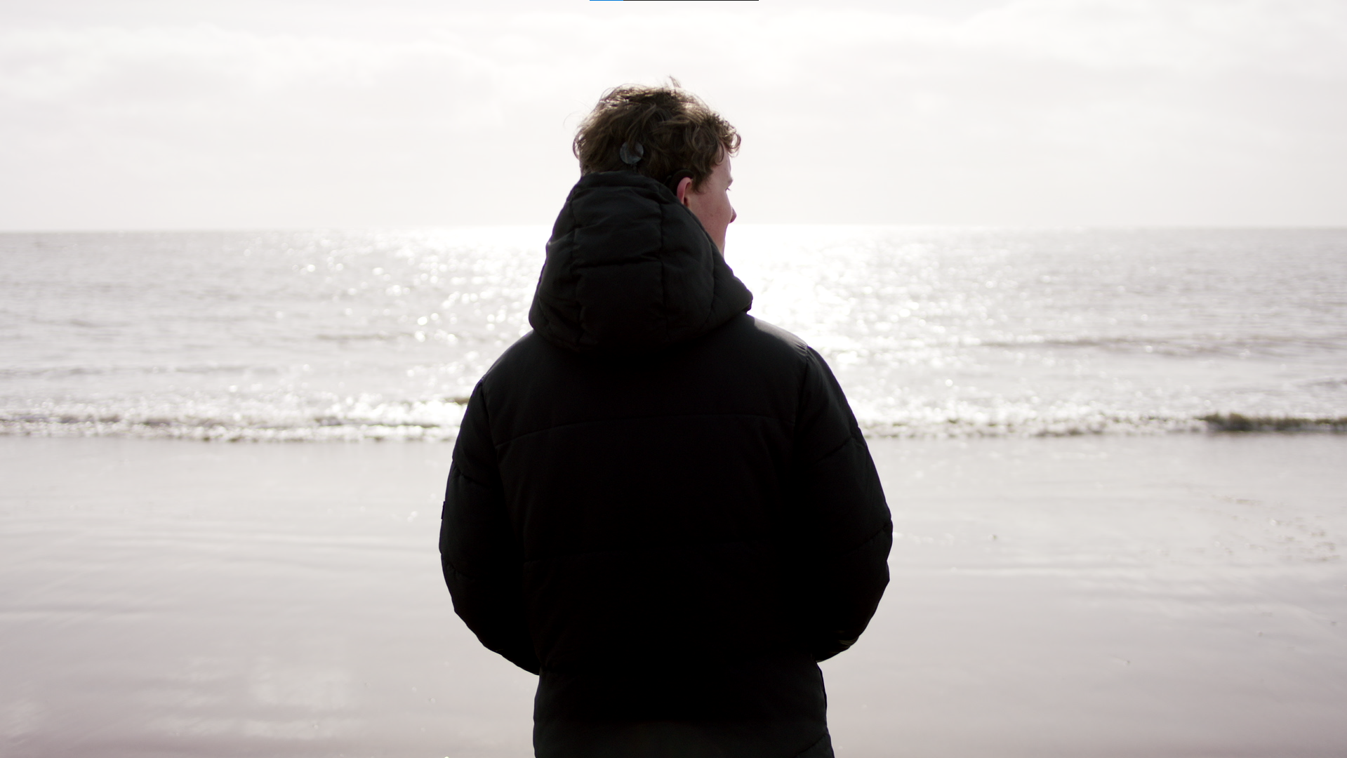Person in black winter coat with hood looking at the ocean on a cloudy day.