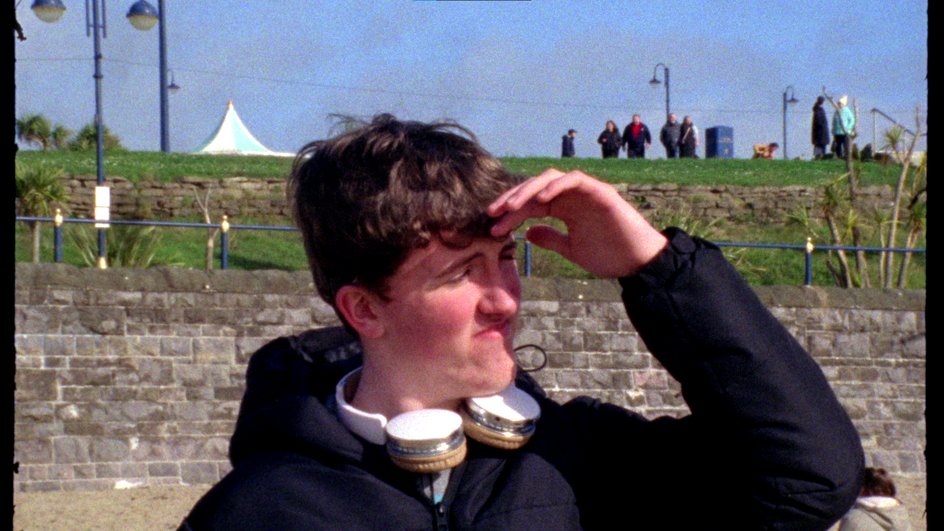 A young man with dark hair looking into the distance, holding his hand above his eyes to shield them from the sunlight, with a cloudy sky, a stone wall, and parked cars behind him.