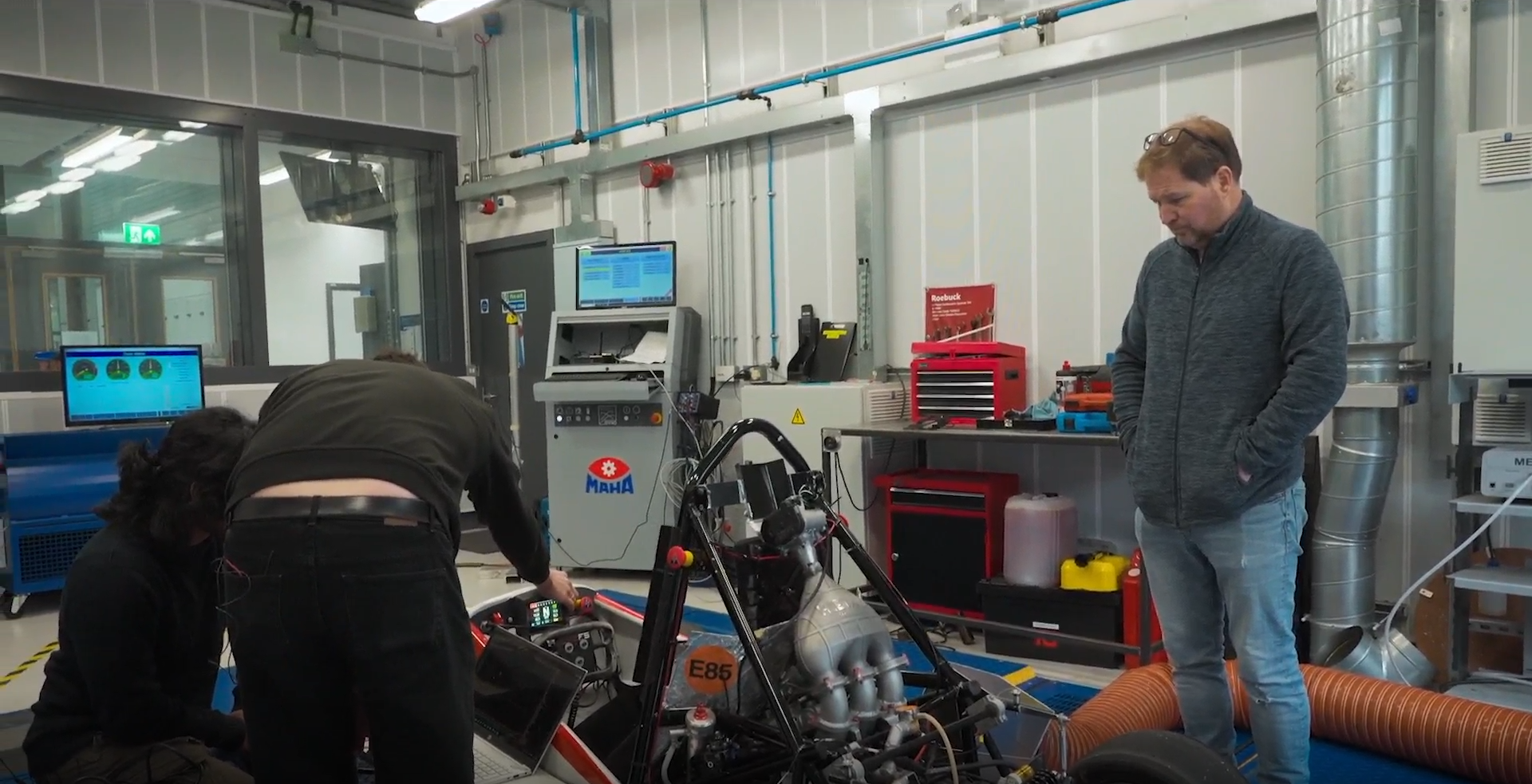 Two men are working on a race car in a workshop, with one man standing and observing and another bent over working on the car's electronic system. The workshop has various tools, machinery, and computer monitors.