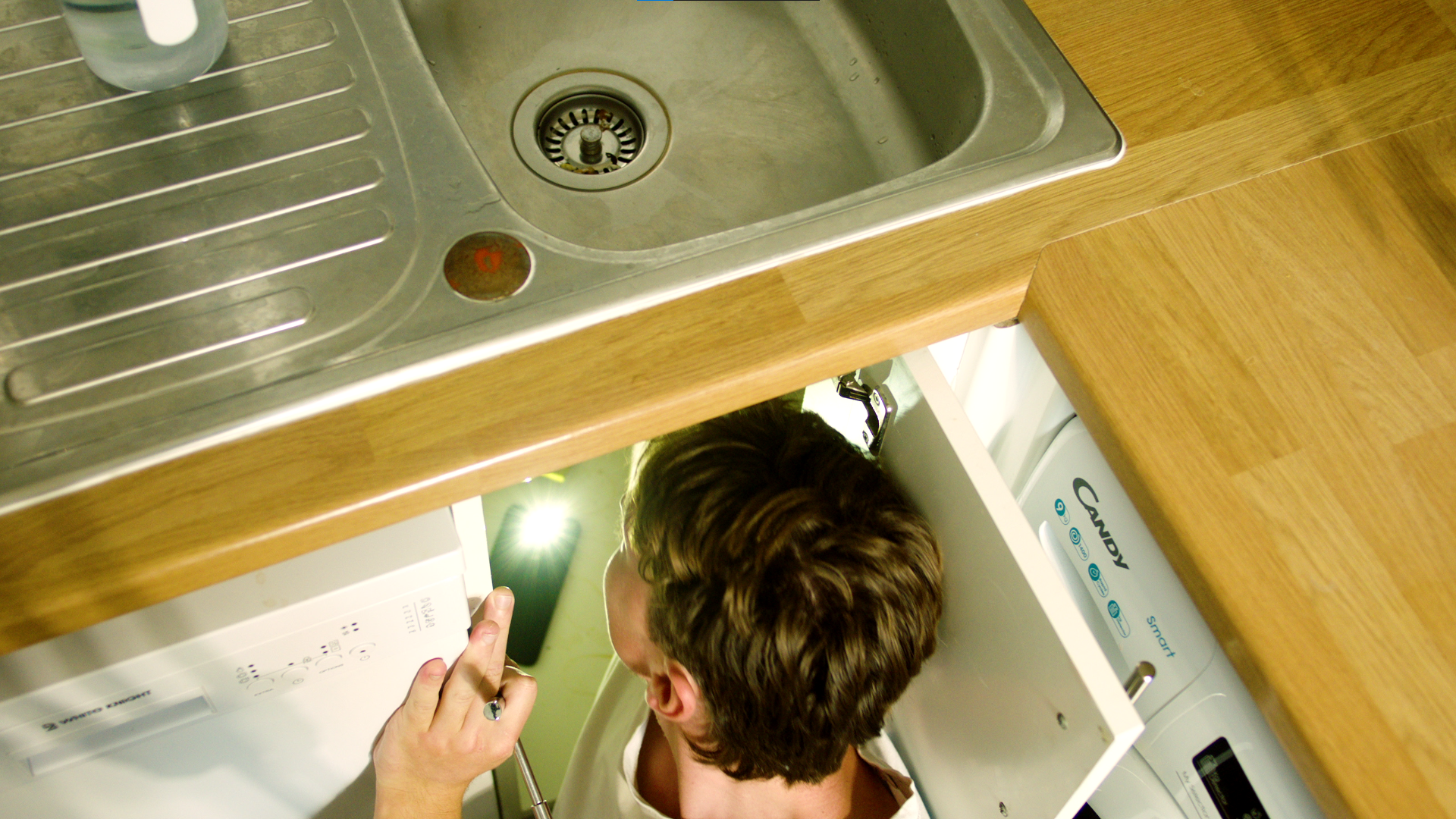 A person repairing or inspecting a washing machine in a laundry room, with a stainless steel kitchen sink and wooden countertop visible.