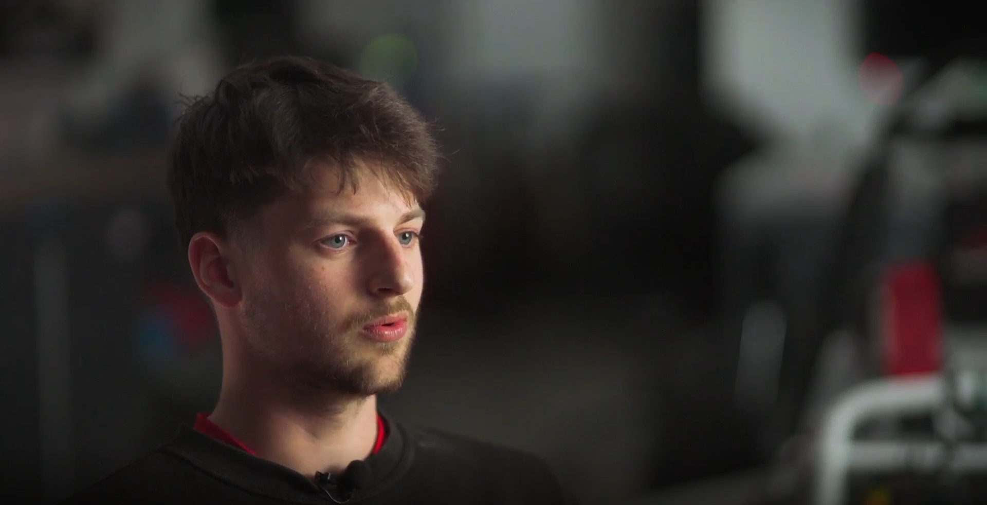 A young man with brown hair and a beard looking thoughtfully at a computer screen in a dark room, possibly in a gym or training facility.