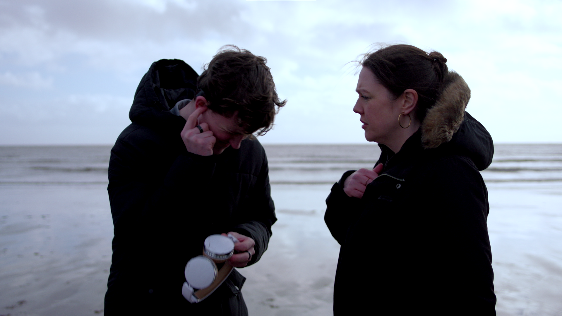 Two women on a beach, one is crying and the other looks concerned, both wearing black jackets.