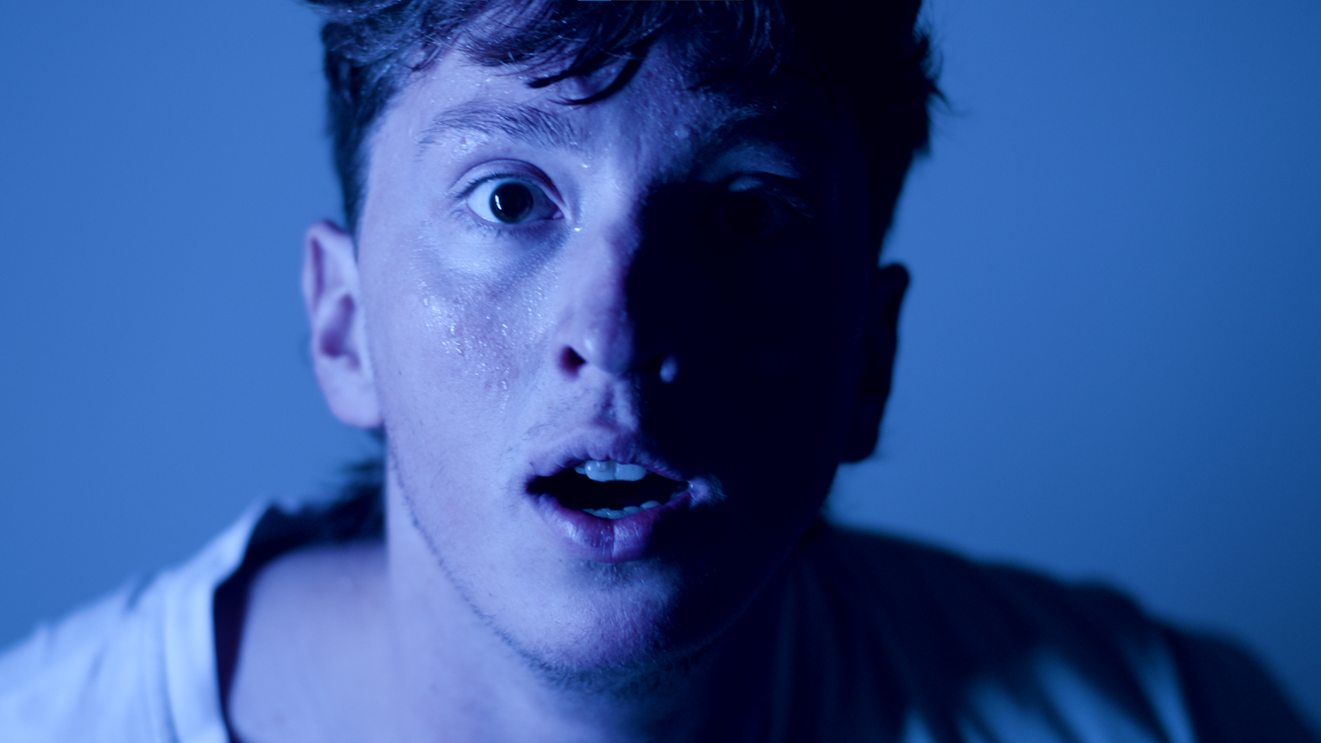 A young man with wet hair and a surprised or shocked expression on his face, illuminated with blue lighting.
