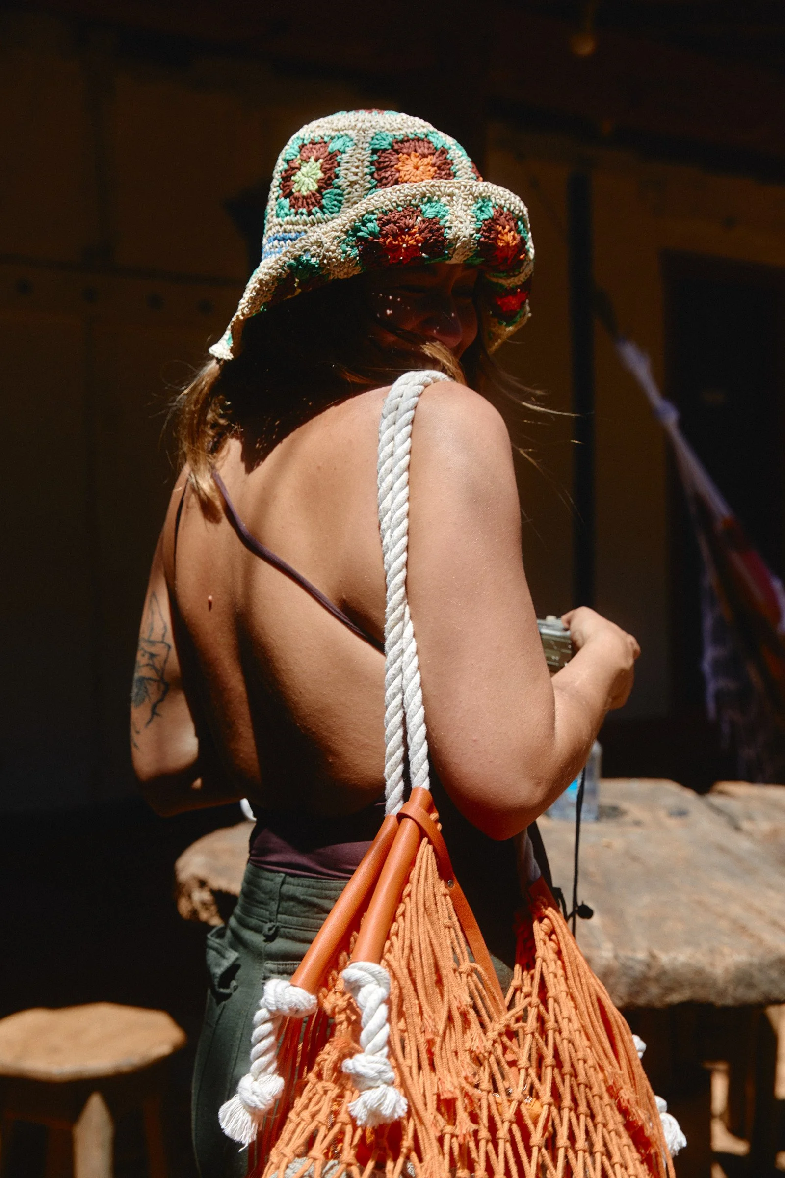 A person with long hair, wearing a colorful crocheted hat with floral patterns, holding an orange woven bag with white knotted rope handles, indoors with dark background.