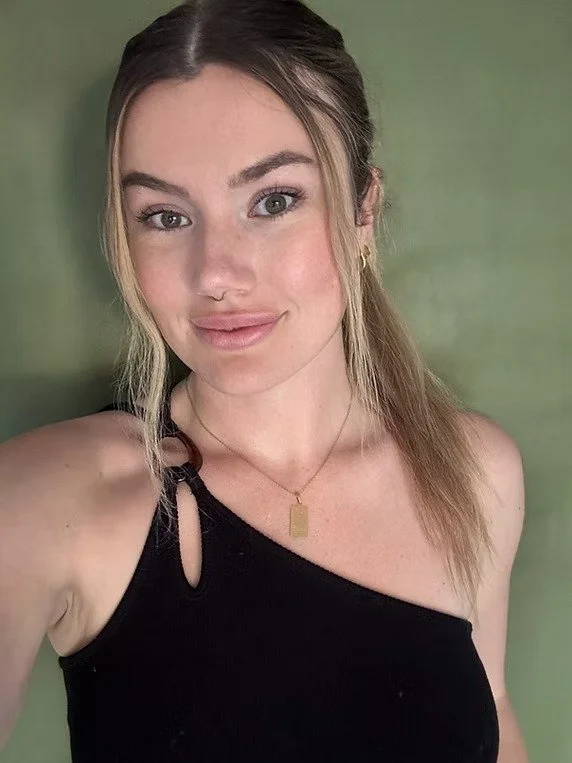 A young woman with light skin and blonde hair taking a selfie. She is wearing a black top with a cutout strap and a gold necklace, and has a slight smile. The background is a green wall.
