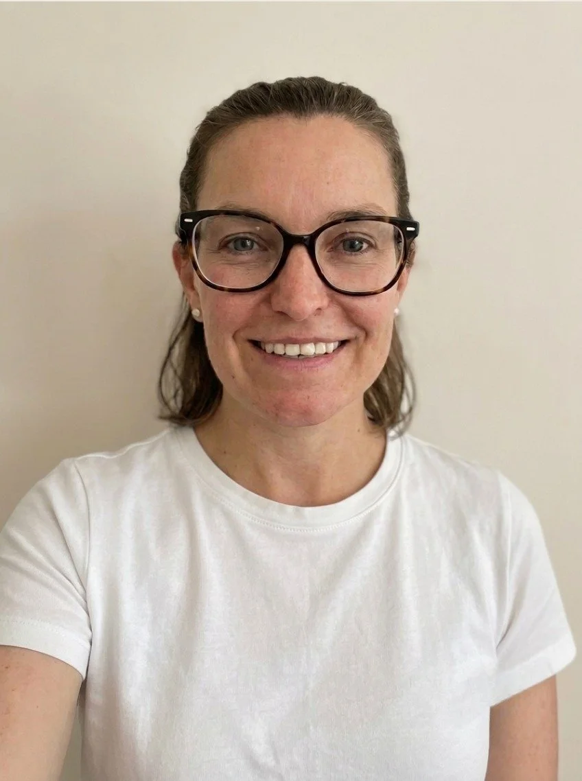 A woman with glasses smiling, wearing a white t-shirt, standing against a plain light-colored wall.