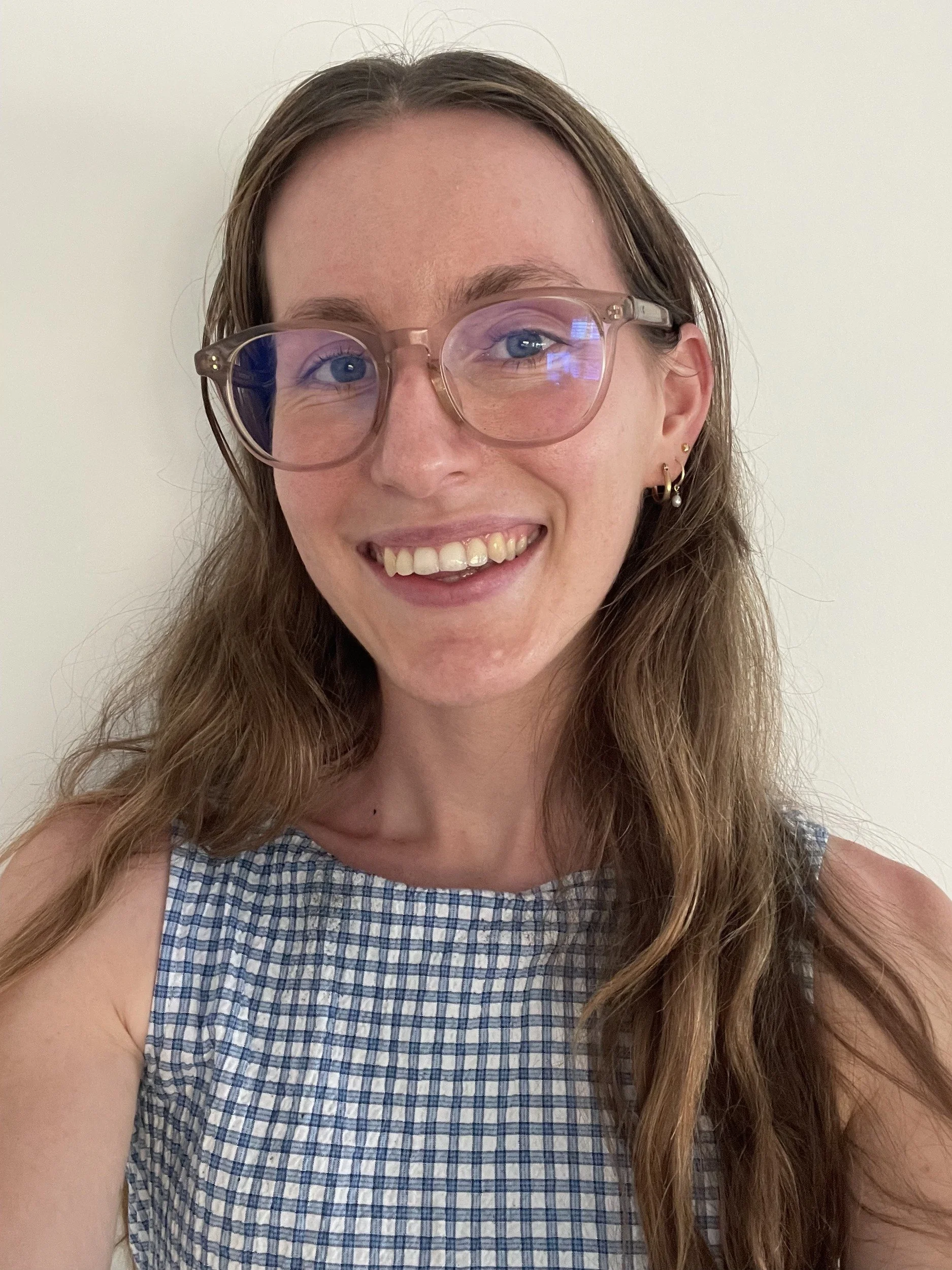 A young woman with long wavy hair, wearing glasses and a sleeveless blue checkered dress, smiling against a plain white background.