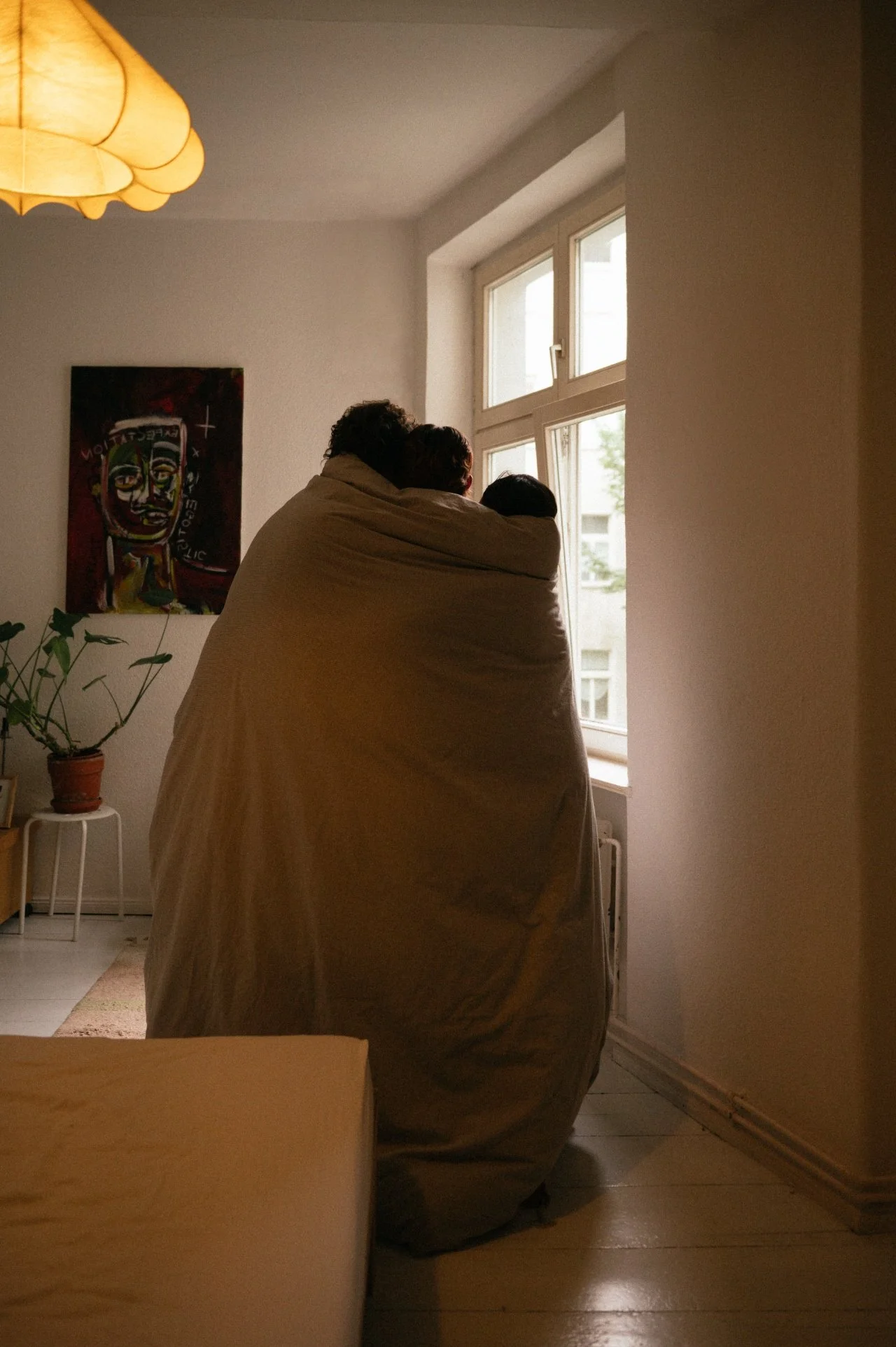 A couple wrapped in a blanket sitting by a window in a room with minimal decor.