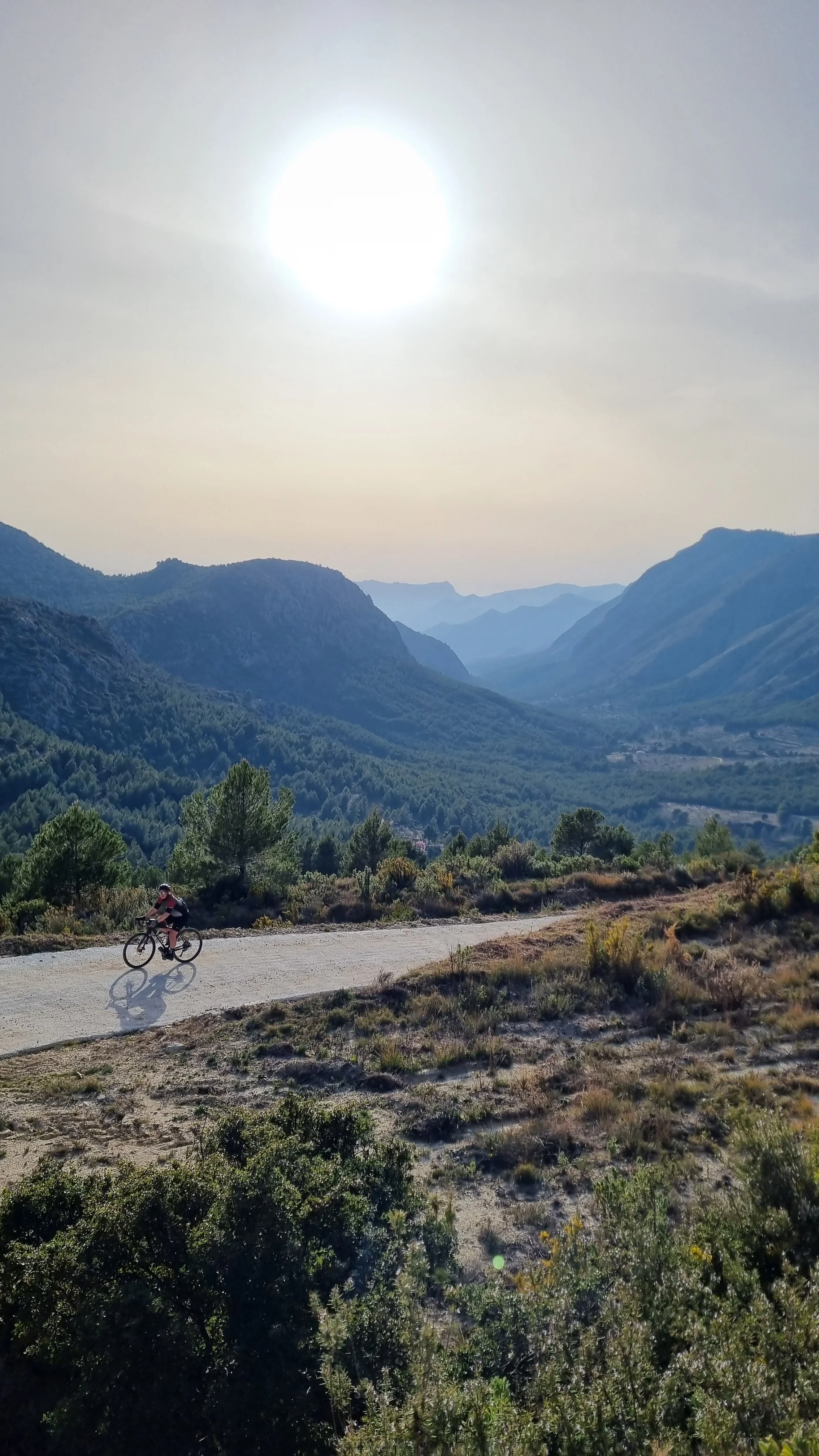 A mountain landscape with a dirt path, green trees, and mountains in the background. A person riding a bicycle is on the path, and the sky is clear with the sun shining brightly.