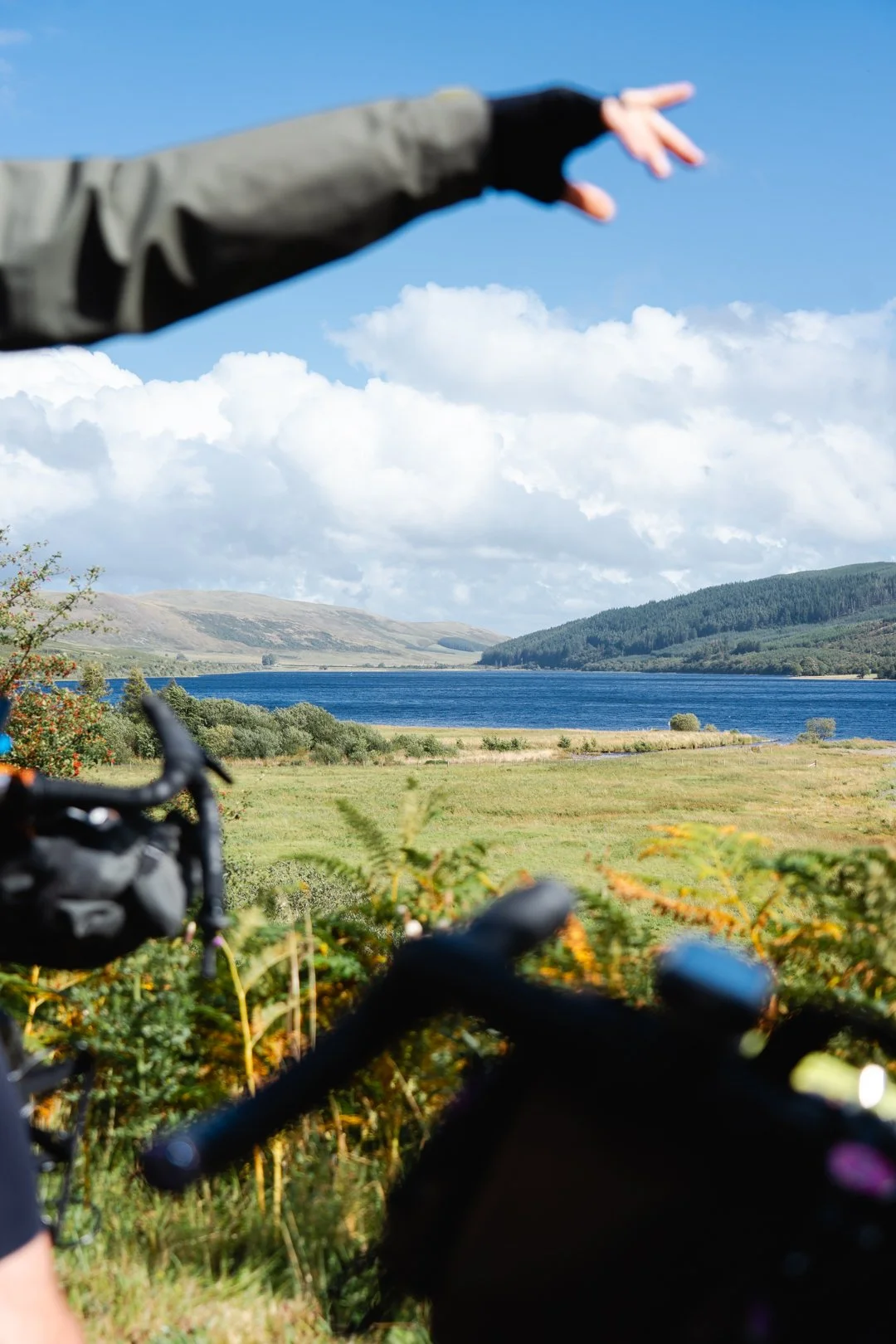 View of a lake and green hills under a blue sky with clouds, seen from behind a bicycle and a person pointing into the distance.