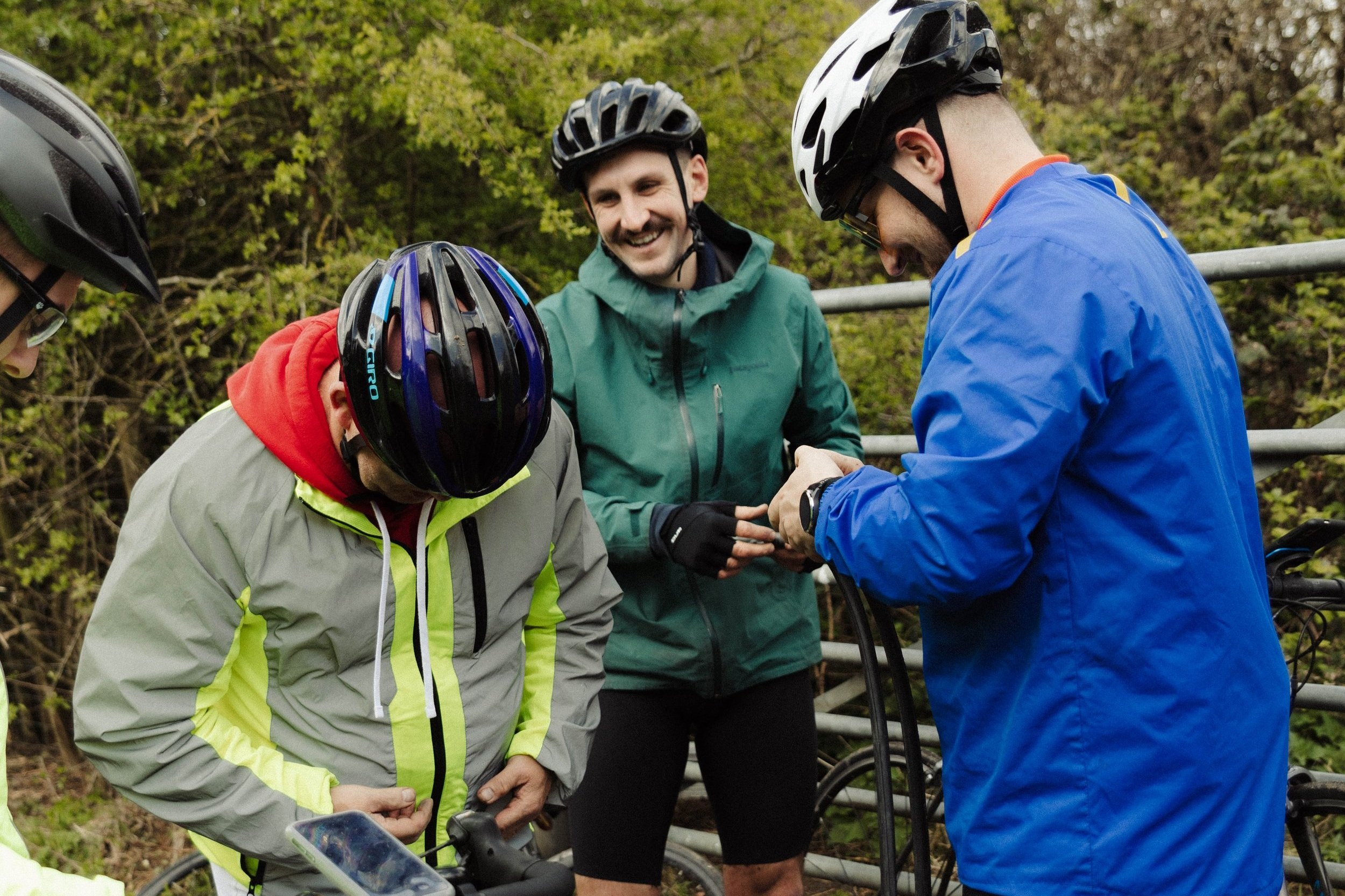 Group of four cyclists, three men and one woman, wearing helmets and outdoor jackets, gathering outdoors in front of green trees, possibly preparing for a bike ride.
