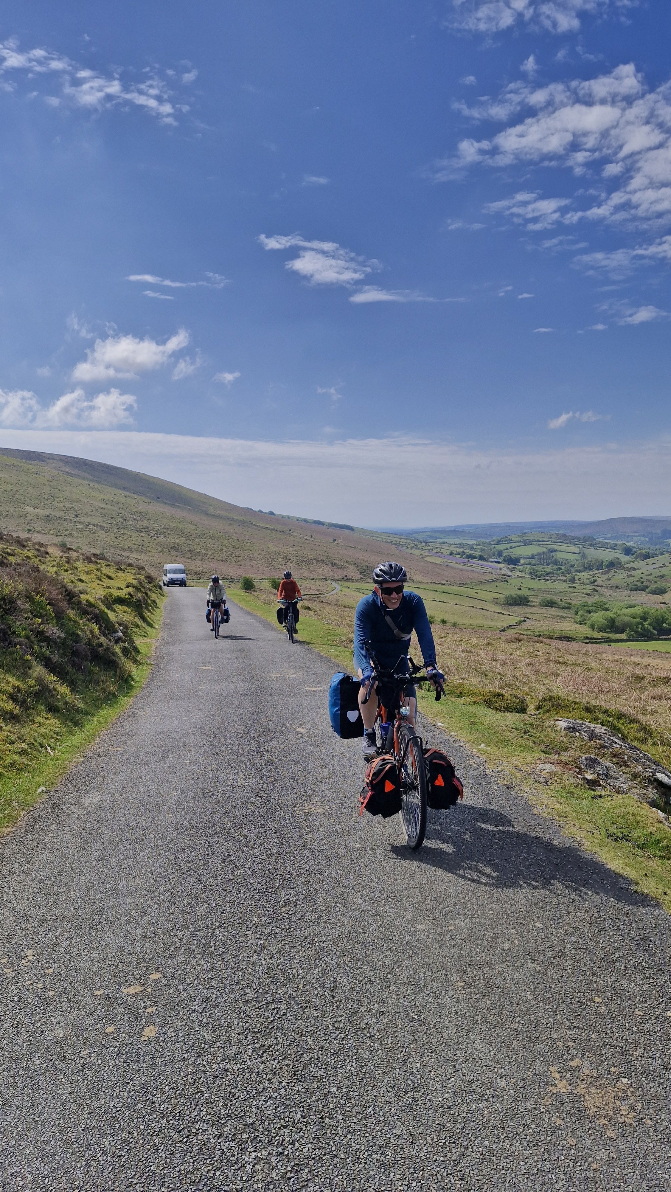 Four cyclists riding on a rural paved road through rolling green hills under a partly cloudy sky.