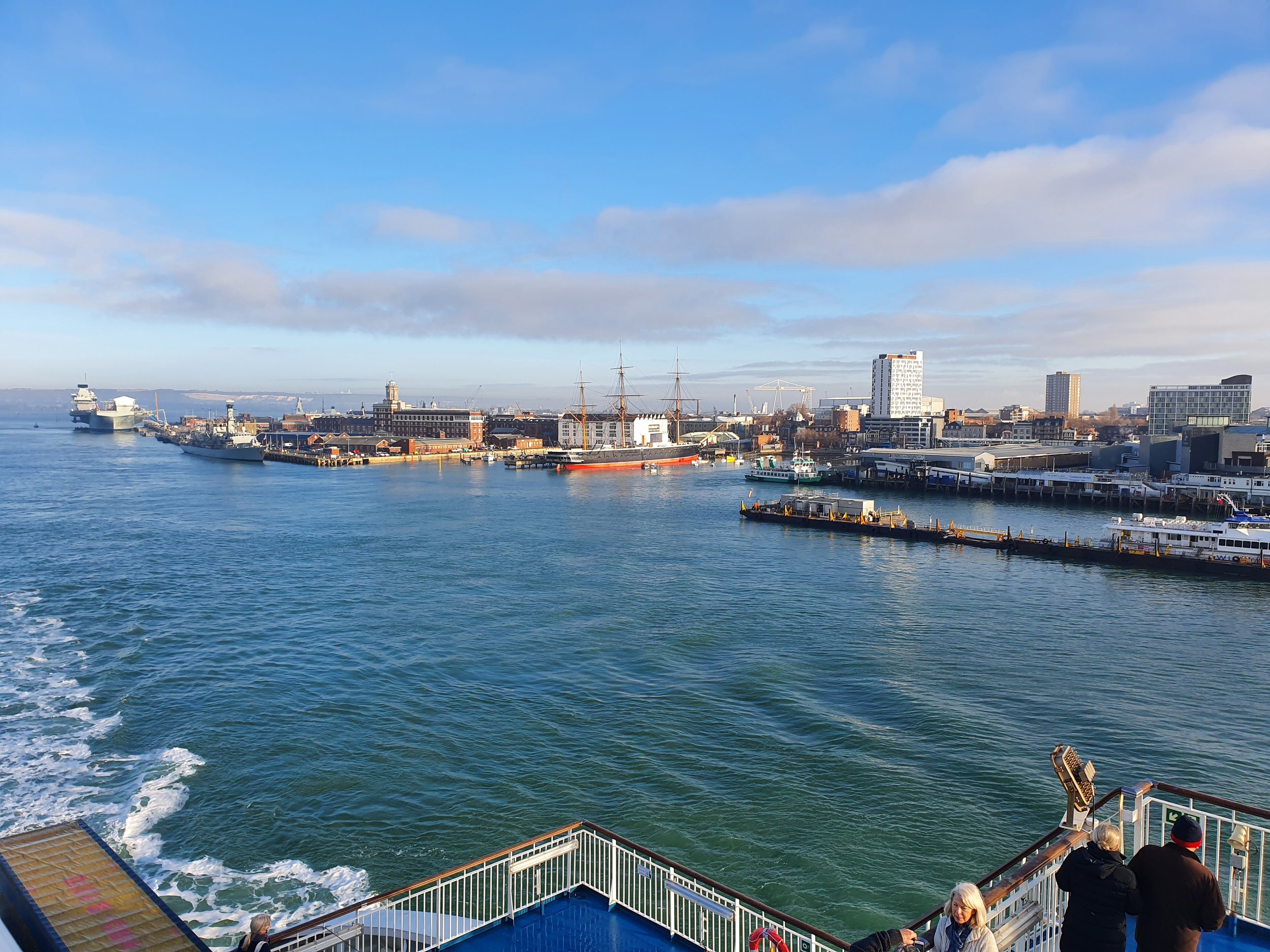 View of a harbor with ships docked, including a large cruise ship and a tall ship with masts, in a city with modern and historic buildings under a partly cloudy sky.