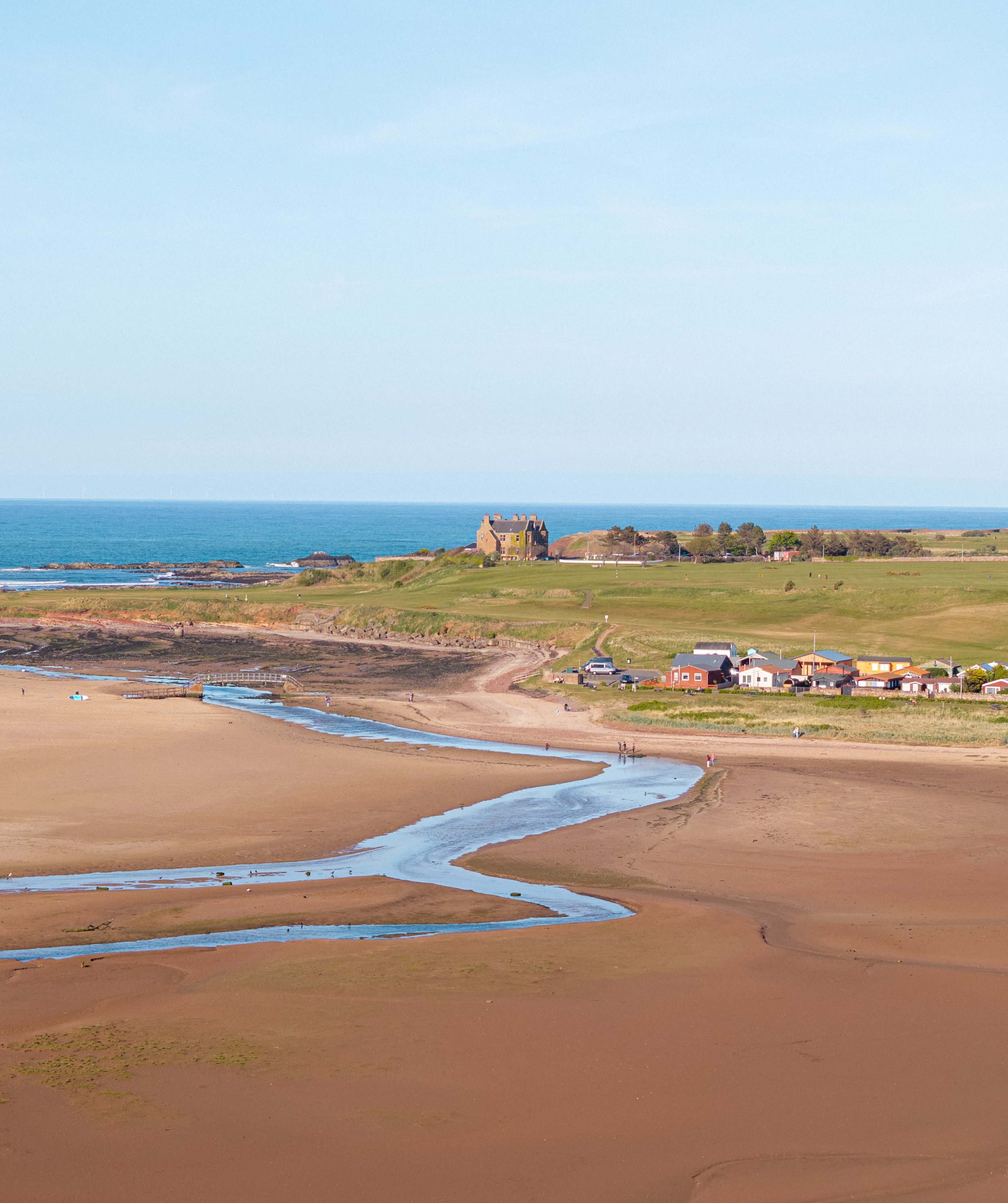 A beach with sandy shores, a small stream, green grass and houses in the background, and the ocean with a clear sky above.