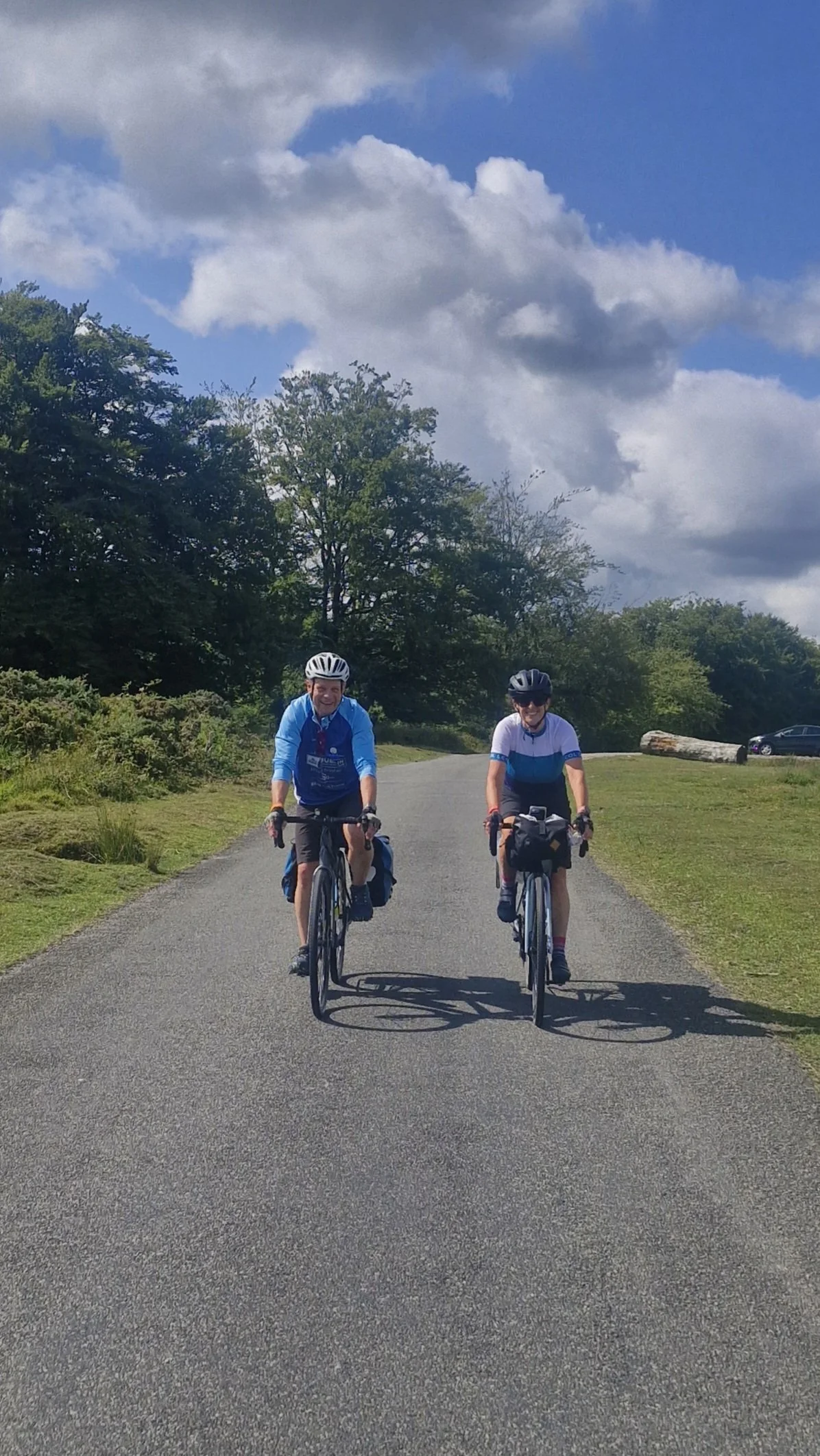Two men cycling on a paved road with trees and a cloudy sky in the background.