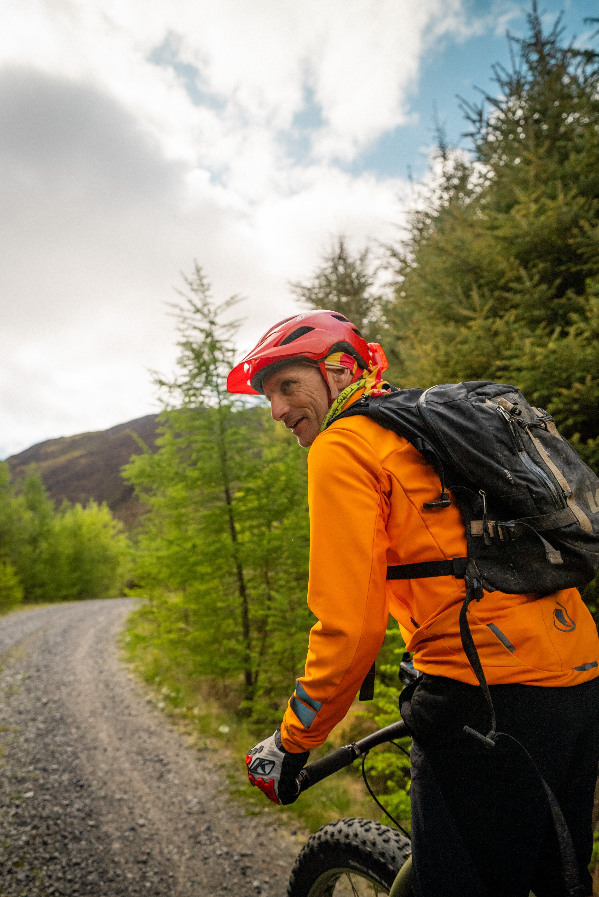 A man riding a mountain bike on a gravel trail surrounded by trees and hills, wearing a red helmet and orange outdoor gear.