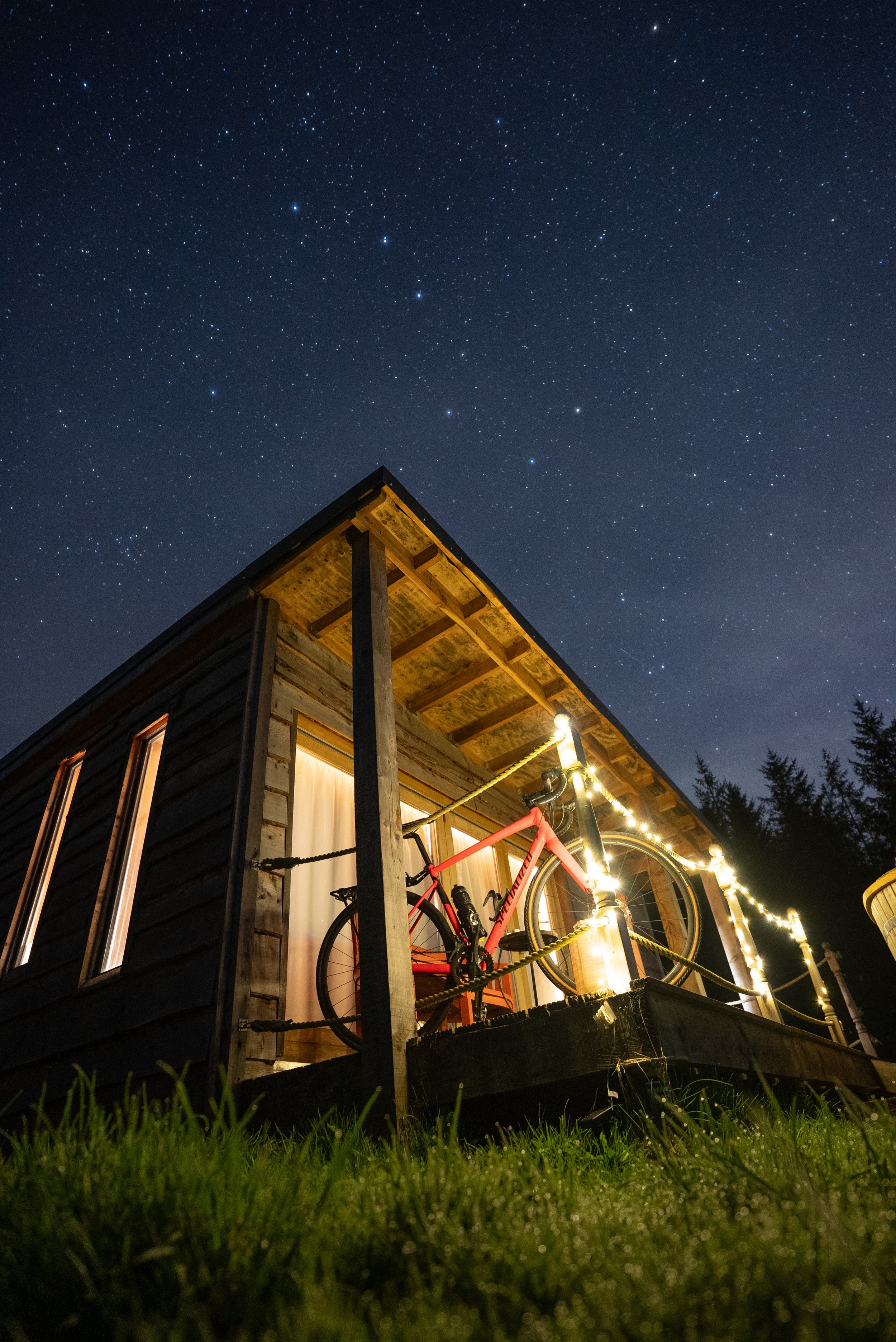 Nighttime view of a wooden balcony with a bicycle decorated with string lights, overlooking a starry sky.