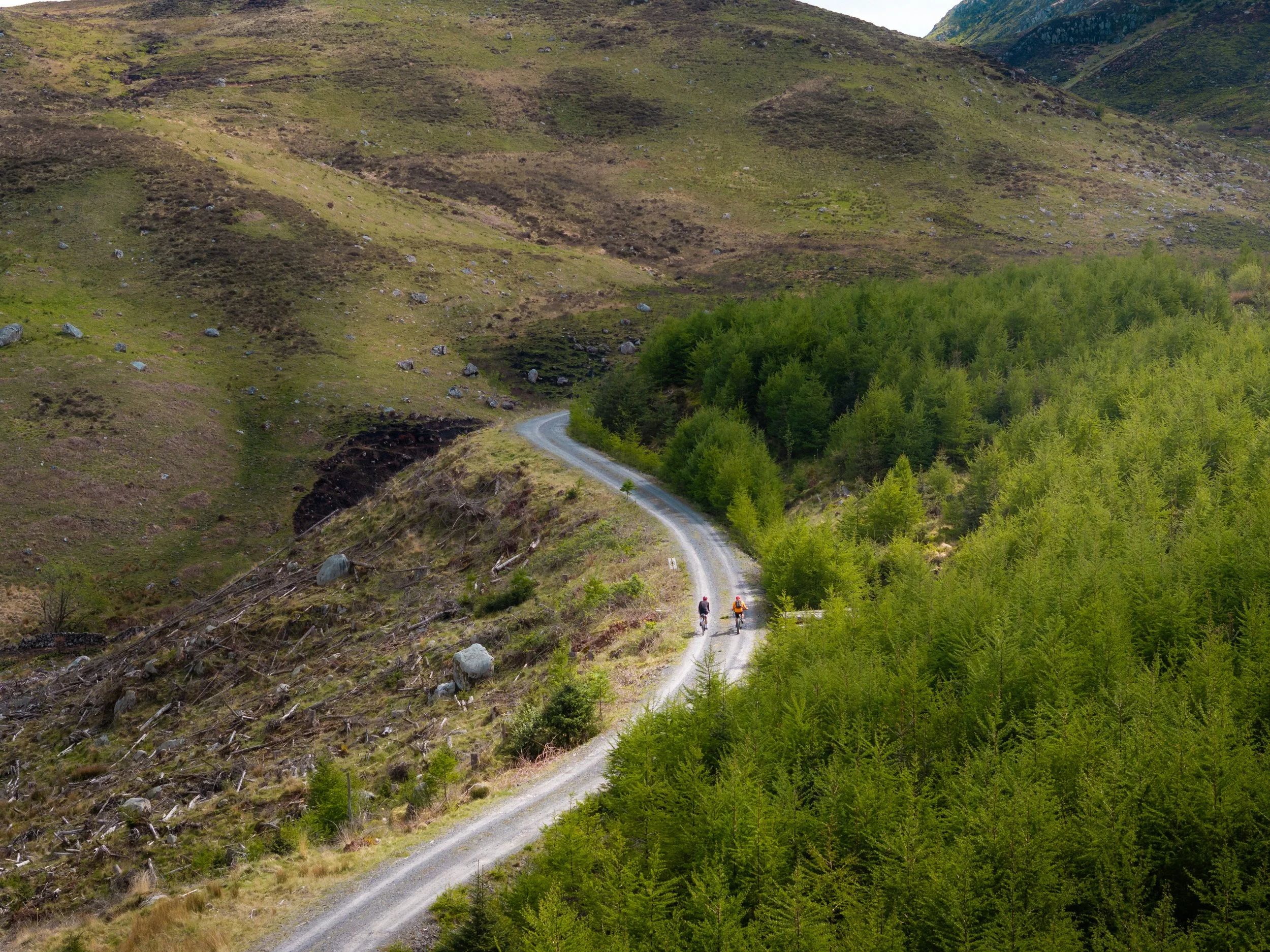 Two cyclists riding on a dirt trail through a lush green valley, surrounded by rolling hills and mountains.