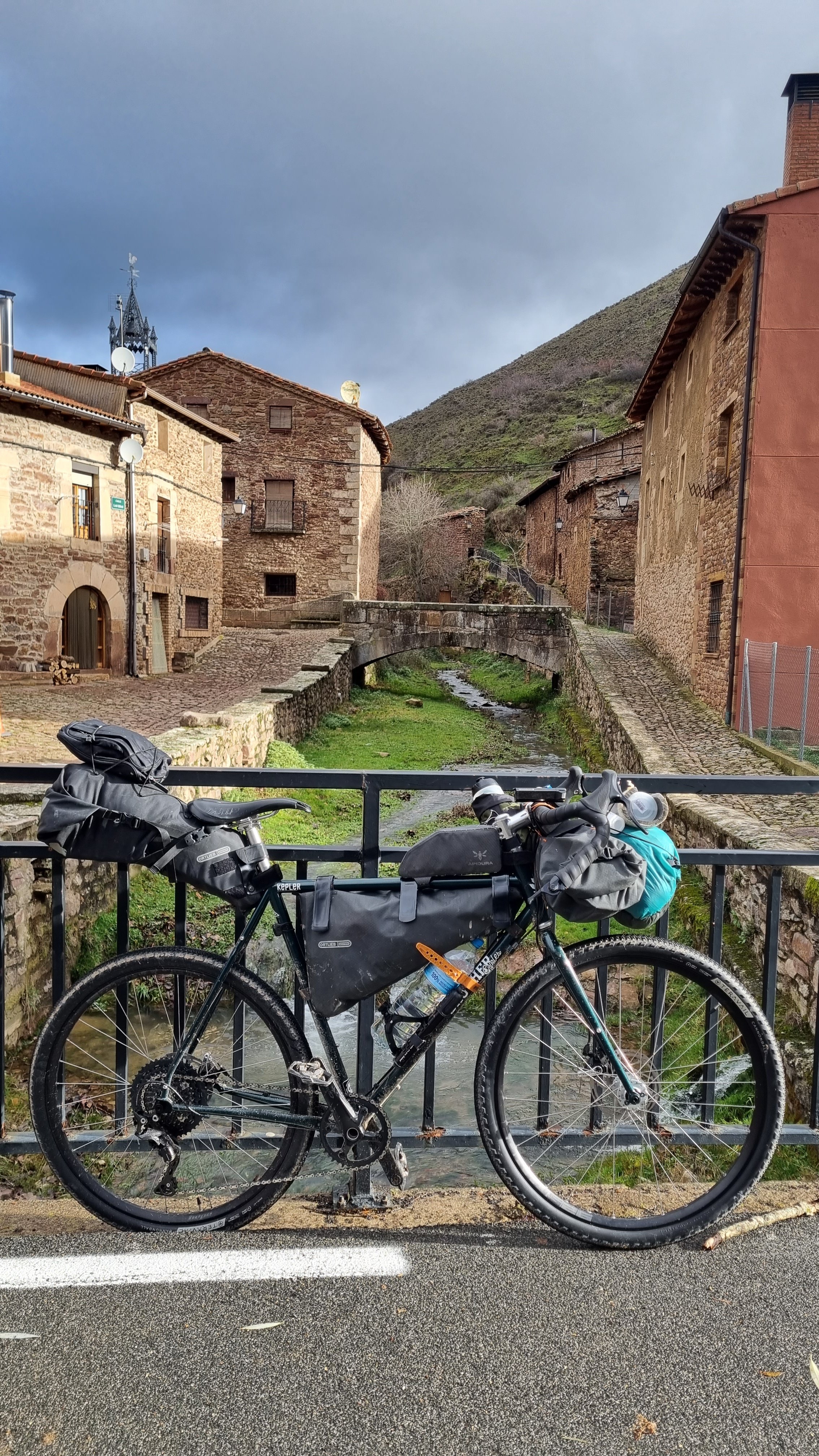 A bicycle with gear and water bottle attached, parked on a bridge over a small stream in a European village with old stone buildings and a mountain in the background.