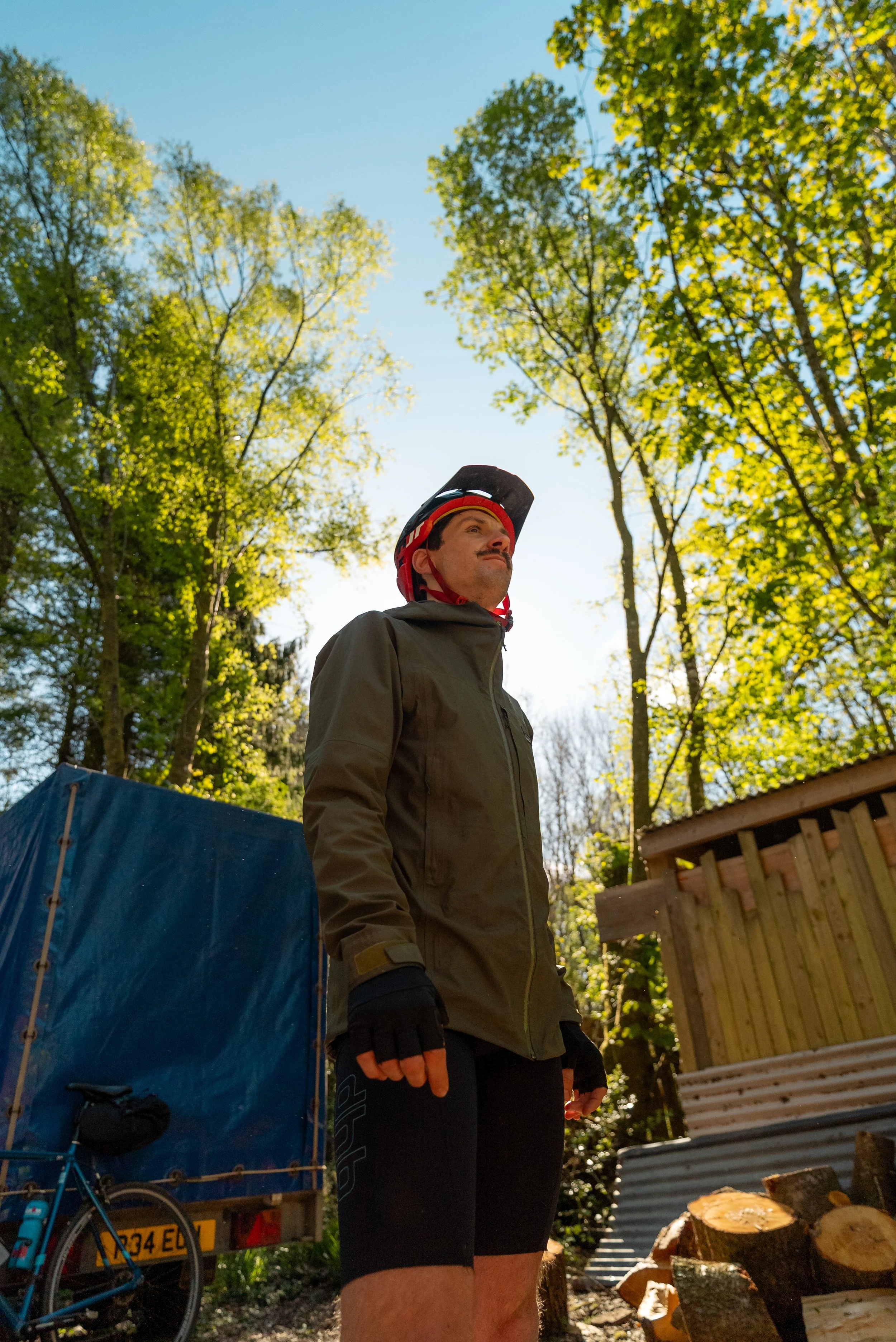 A man wearing a bicycle helmet and outdoor clothing standing outdoors with trees and a wooden structure in the background.