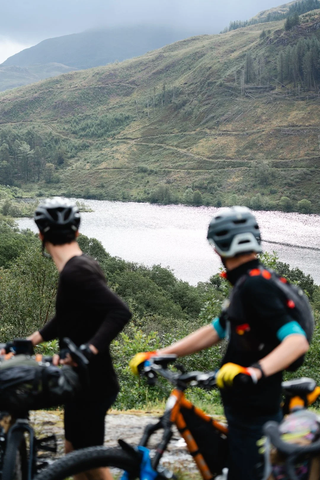 Two cyclists wearing helmets and cycling gear riding mountain bikes past green trees with a lake and hills in the background.