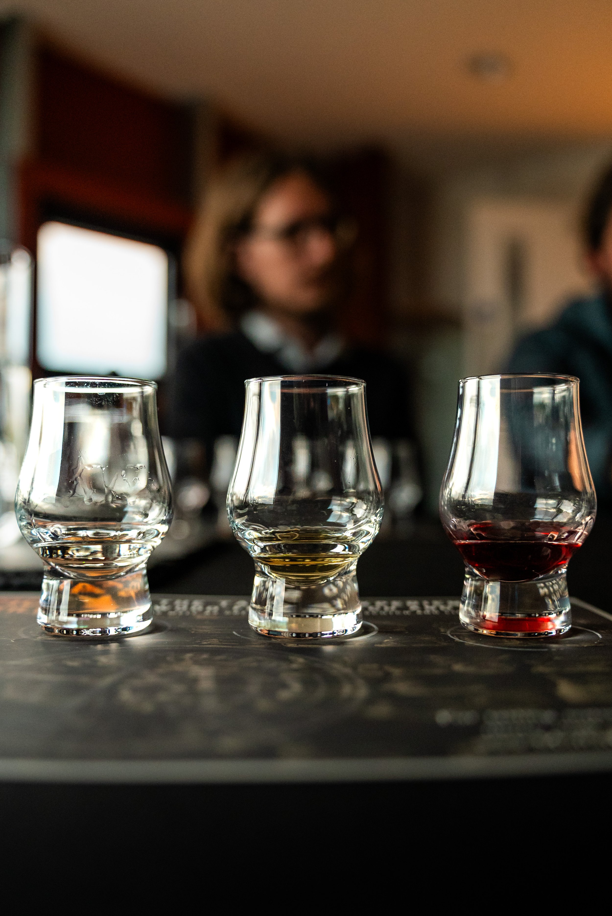 Four glasses of varying liquids on a countertop, with a woman in the background observing them, blurred and out of focus.