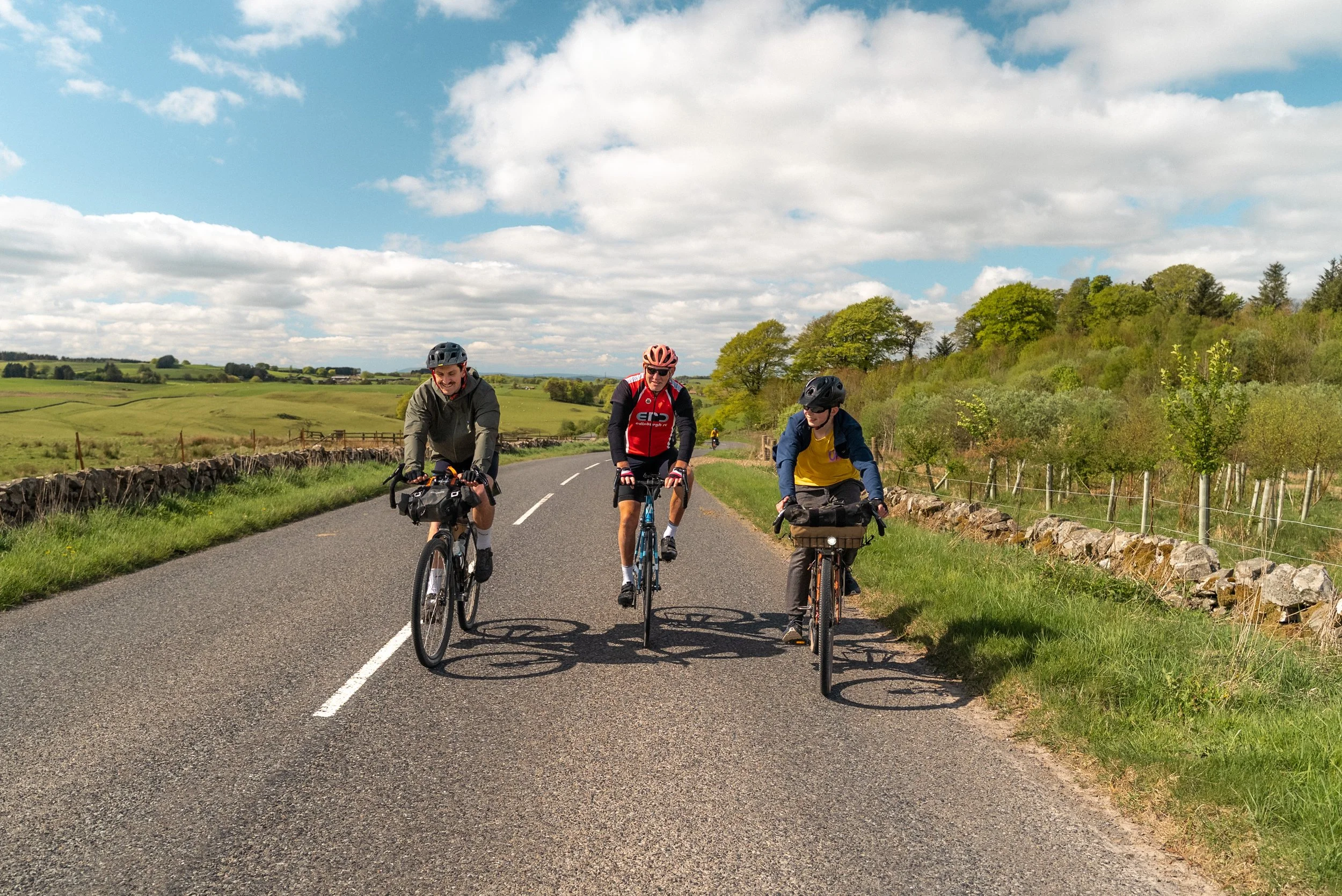 Three cyclists riding on a rural road with green fields and trees under a partly cloudy sky.