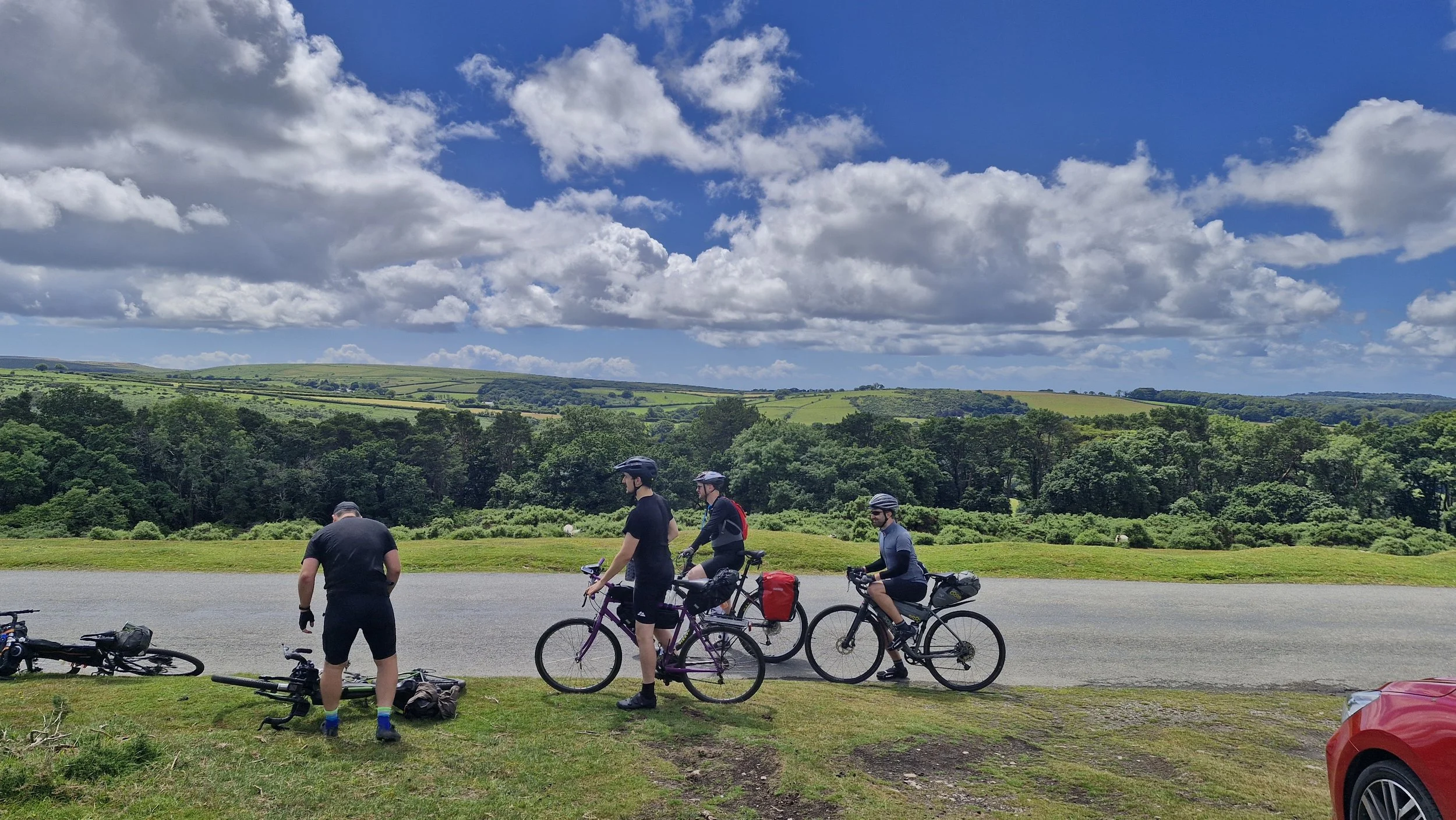 Group of cyclists taking a break on the side of a rural road, with green fields and hills under a partly cloudy sky in the background.