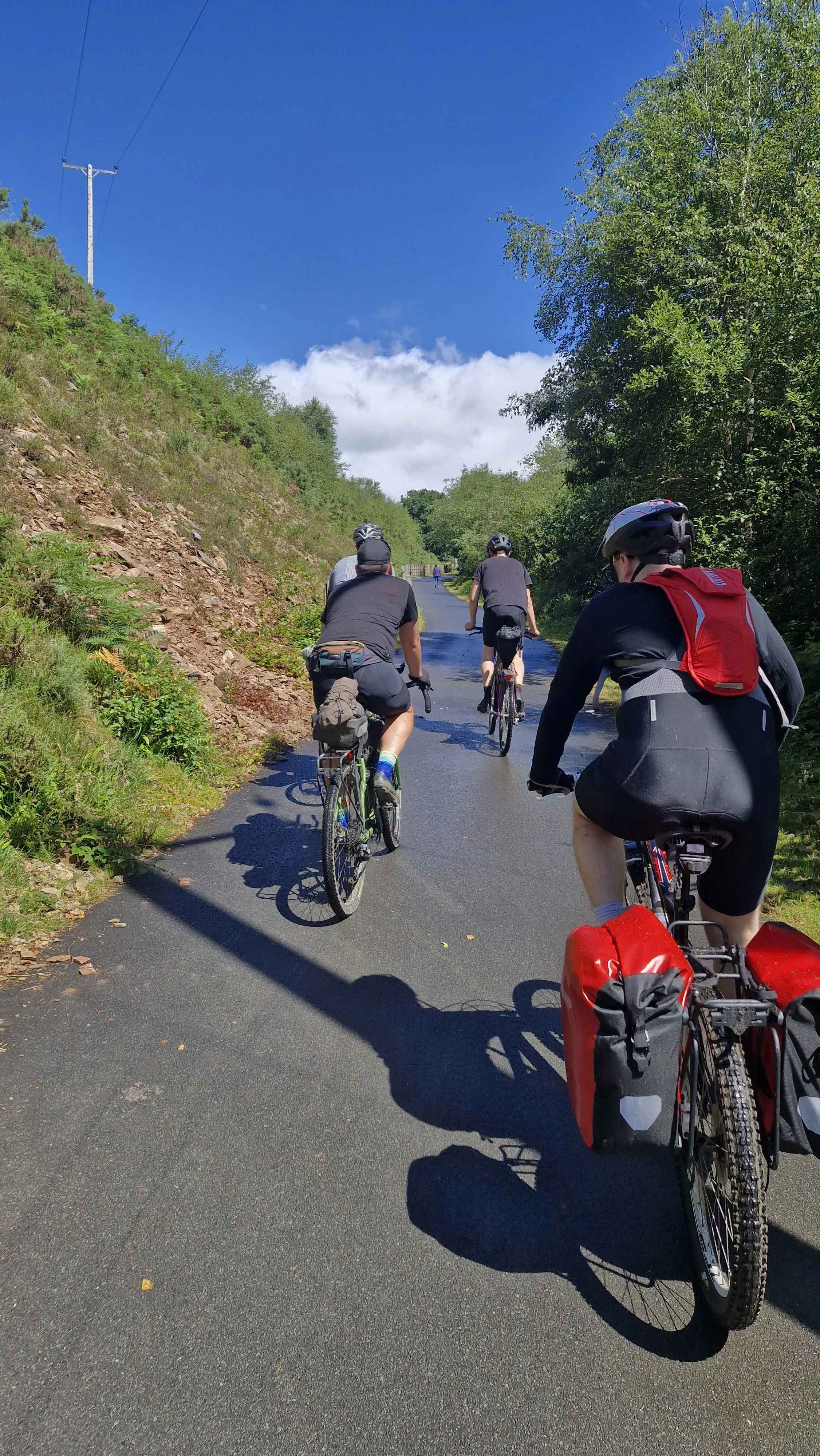 A group of people riding bicycles on a scenic, paved path through a green, hilly area with trees, under a bright blue sky with clouds.