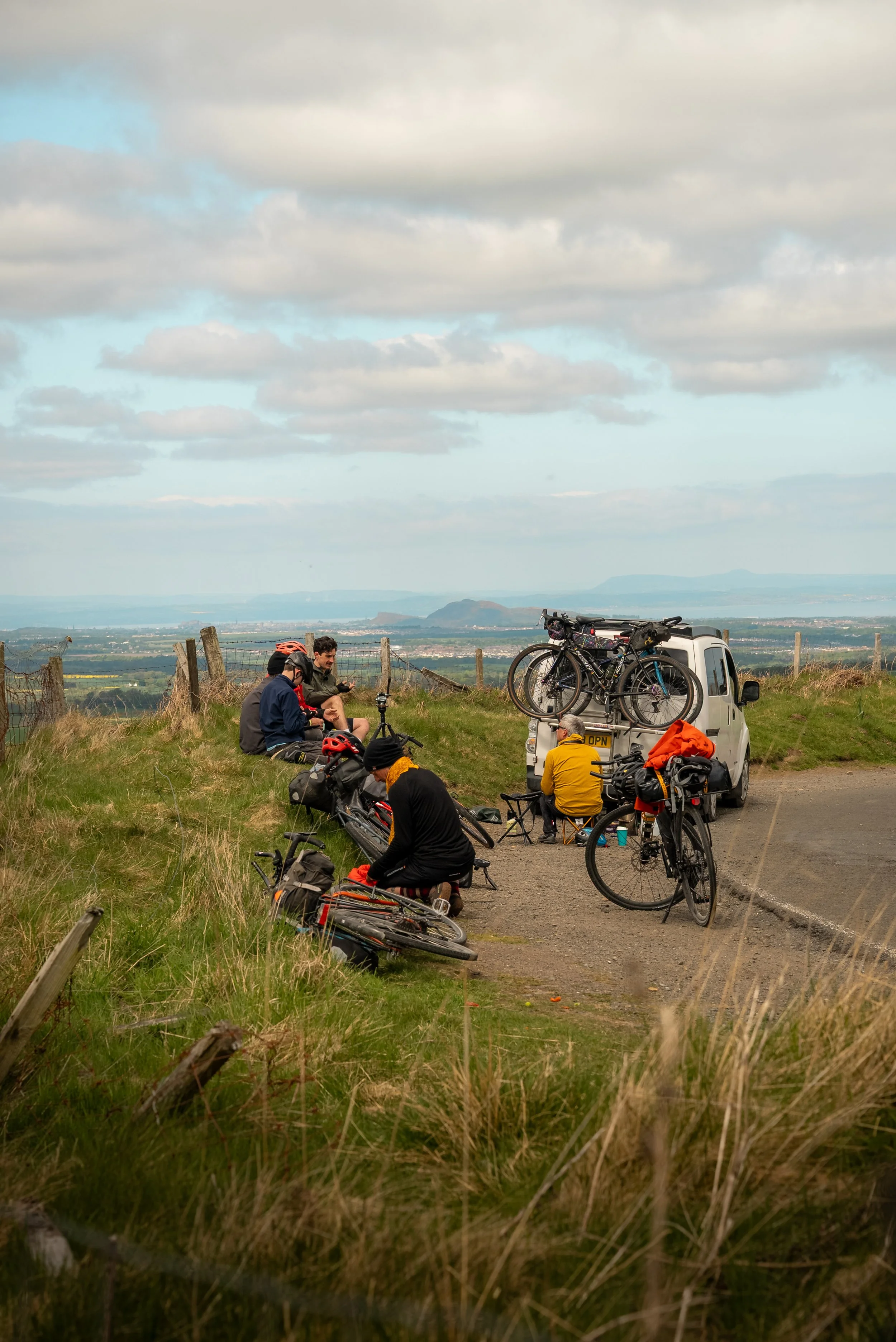 Group of cyclists resting and chatting near a small white van with bikes mounted on top, on a grassy hillside with a scenic view of distant hills and cloudy sky.