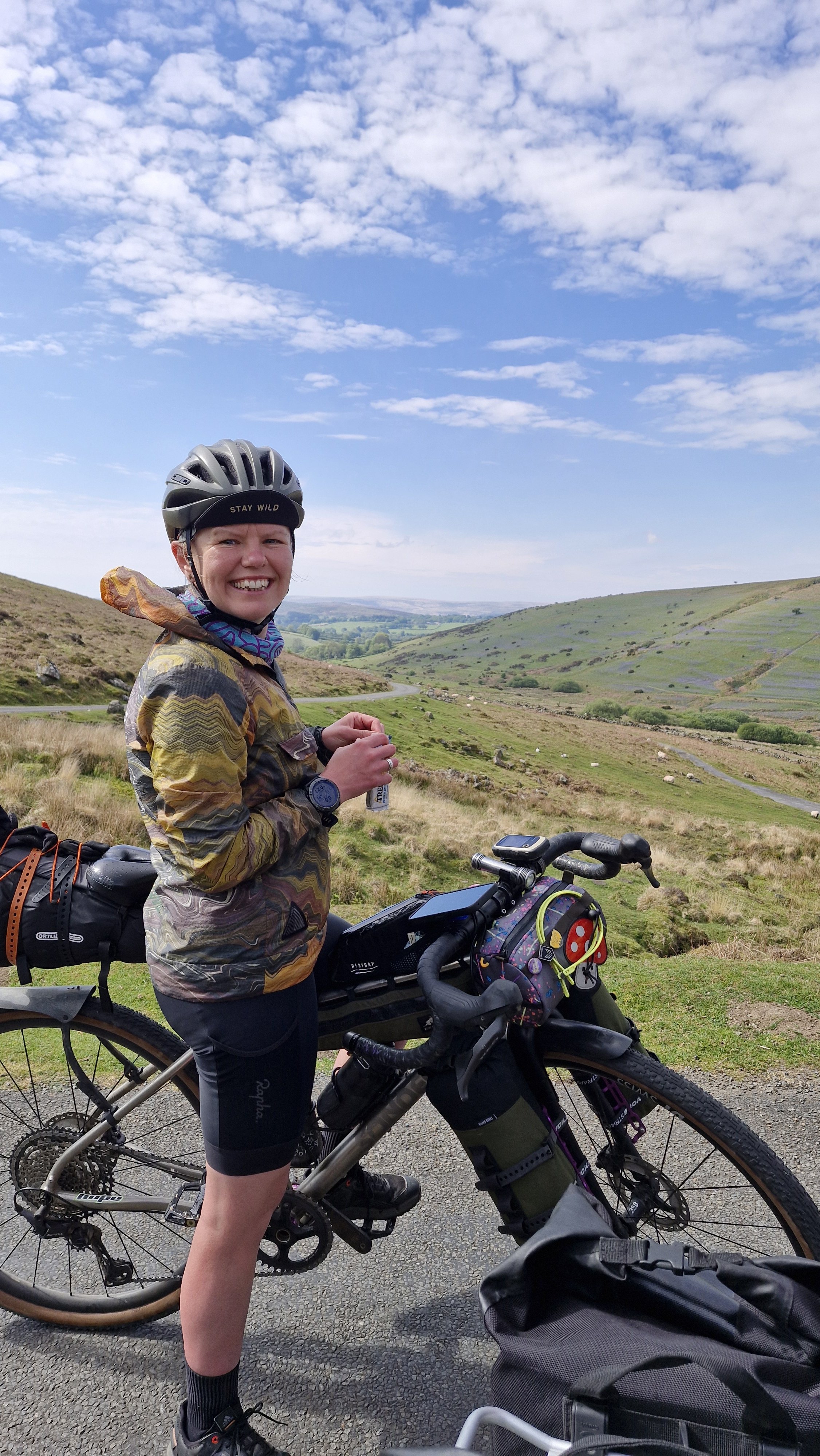 A smiling woman dressed in cycling gear standing next to her mountain bike in a scenic open countryside with rolling green hills and a partly cloudy sky.