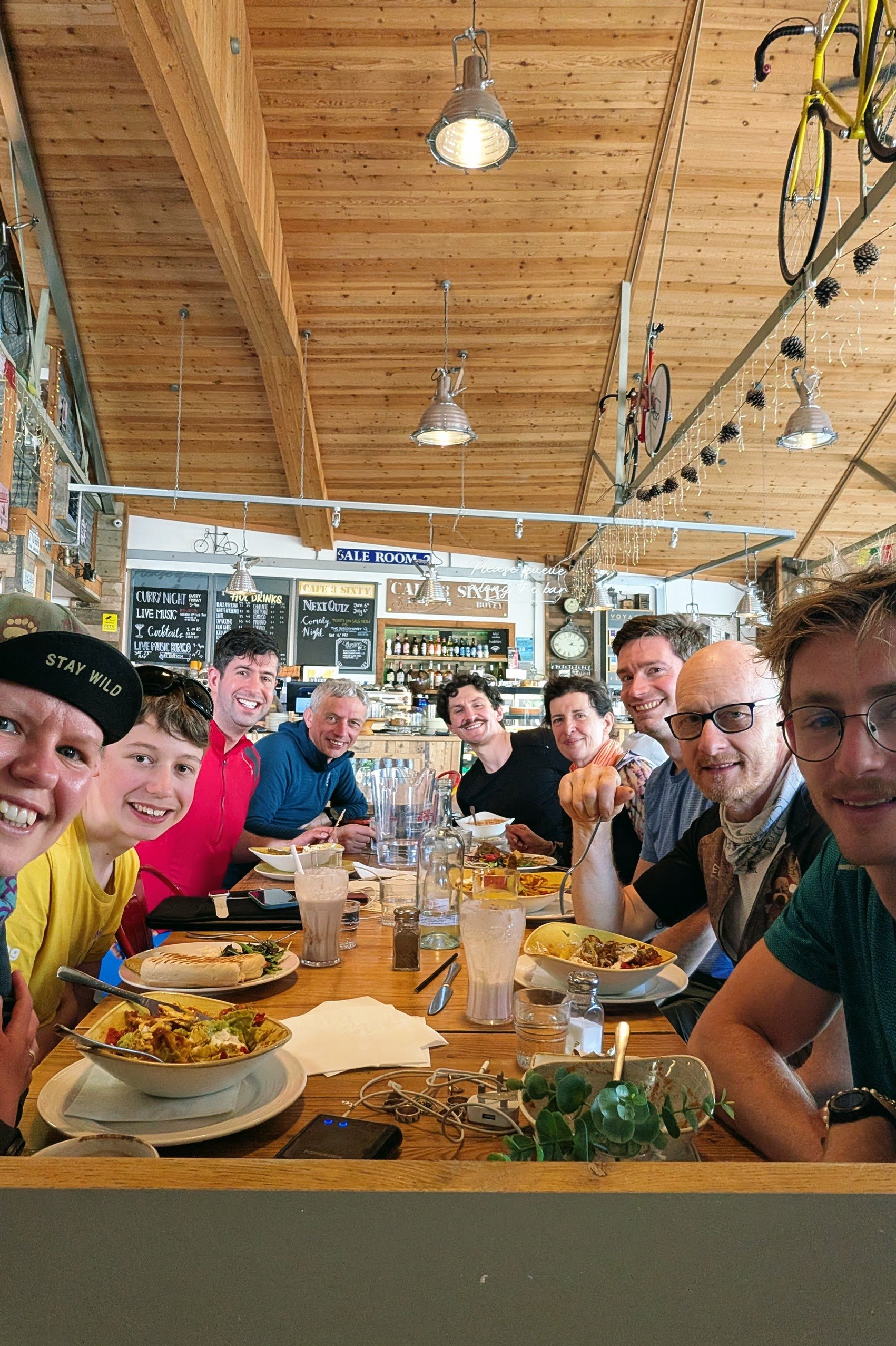 A group of people sitting around a wooden table in a cozy restaurant with wooden ceilings. They are smiling, enjoying food and drinks. There are various dishes and glasses on the table, and some electronics are visible. The restaurant has hanging lights and decor including bikes and pine cones.