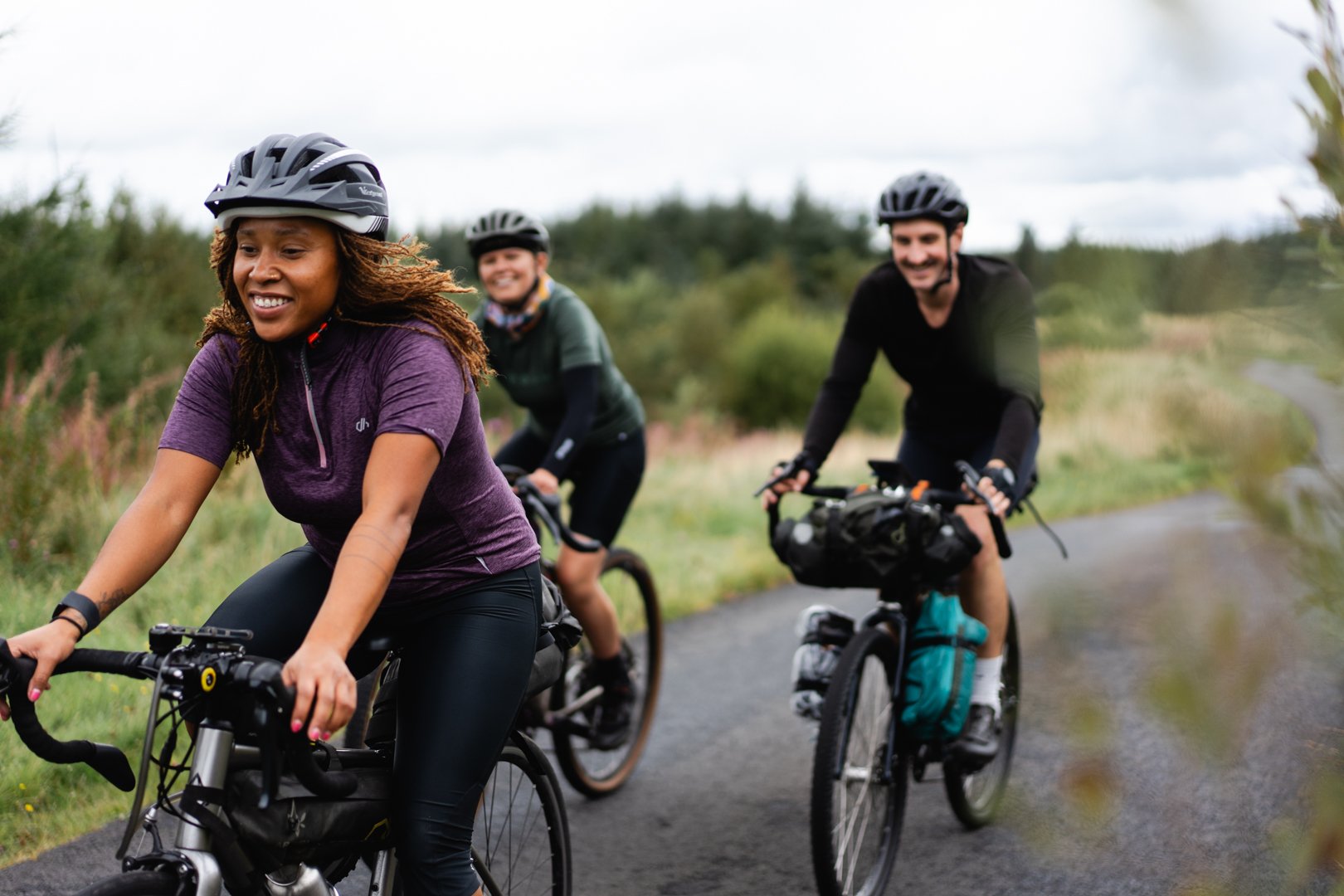 Three people riding bicycles on a paved trail through a natural, green landscape during overcast weather. The woman in front is smiling and wearing a purple shirt and helmet, with two men following behind, also smiling and wearing helmets.