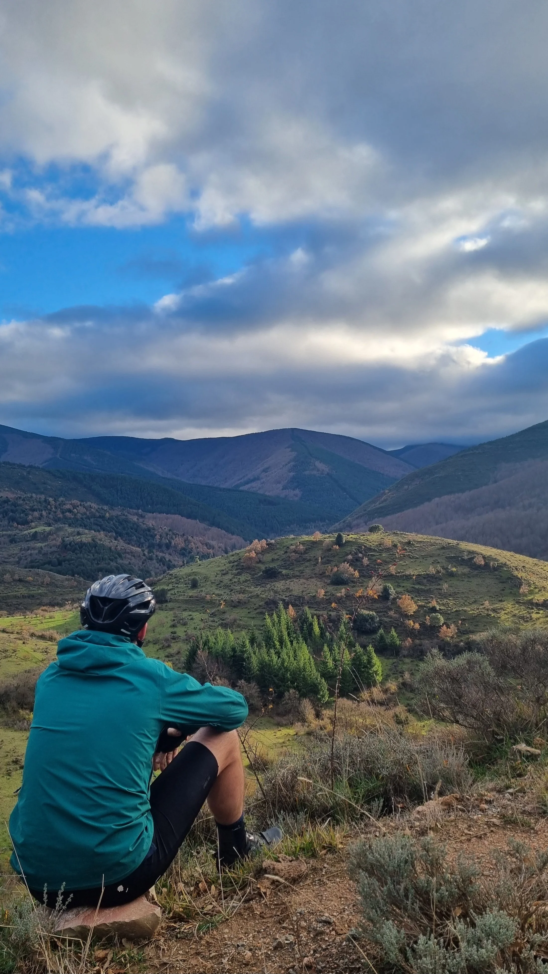 A person in a teal jacket and black helmet sitting on a rock, overlooking a scenic landscape of rolling hills, green trees, and mountain ranges under a partly cloudy sky.